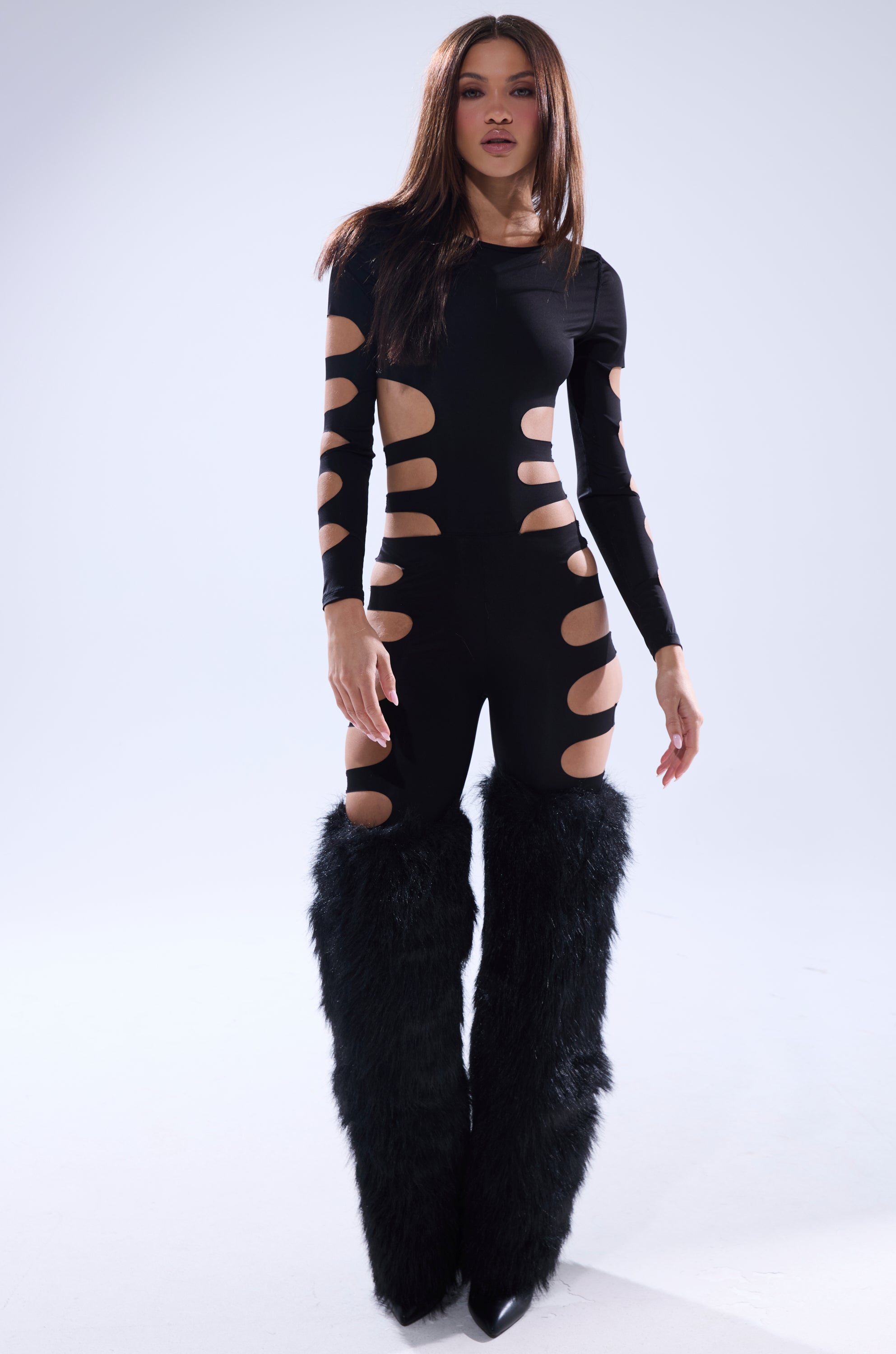 A person stands confidently in the OUT OF HIS LEAGUE JUMPSUIT, a black jumpsuit with bold cutouts along the arms, torso, and legs, paired with large furry black leg warmers against a plain light background.