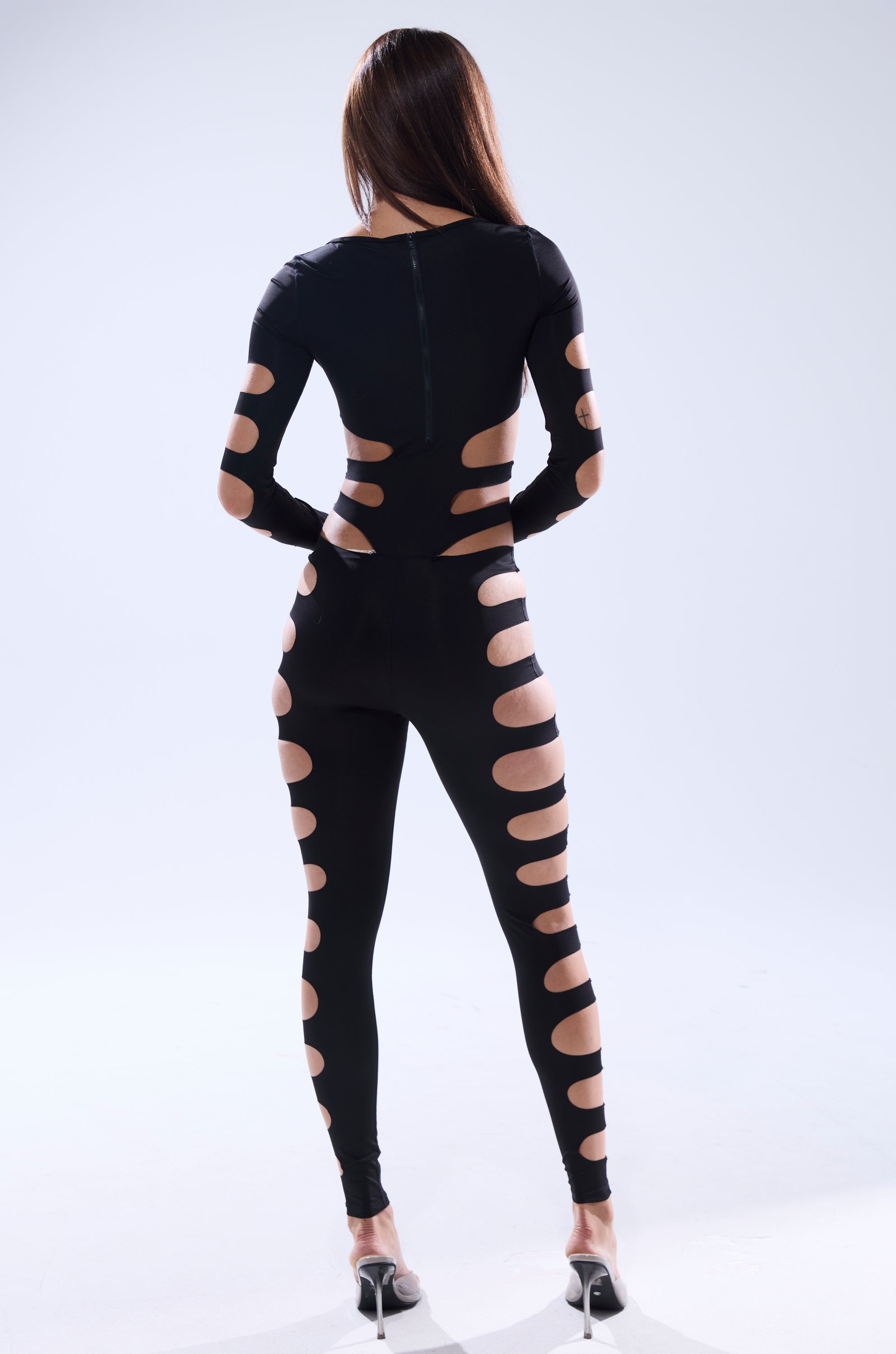 A woman faces away, showcasing the OUT OF HIS LEAGUE JUMPSUIT—a black cutout design with large circular openings along the arms, sides, and legs. She has long straight hair and wears high heels against a plain light background.