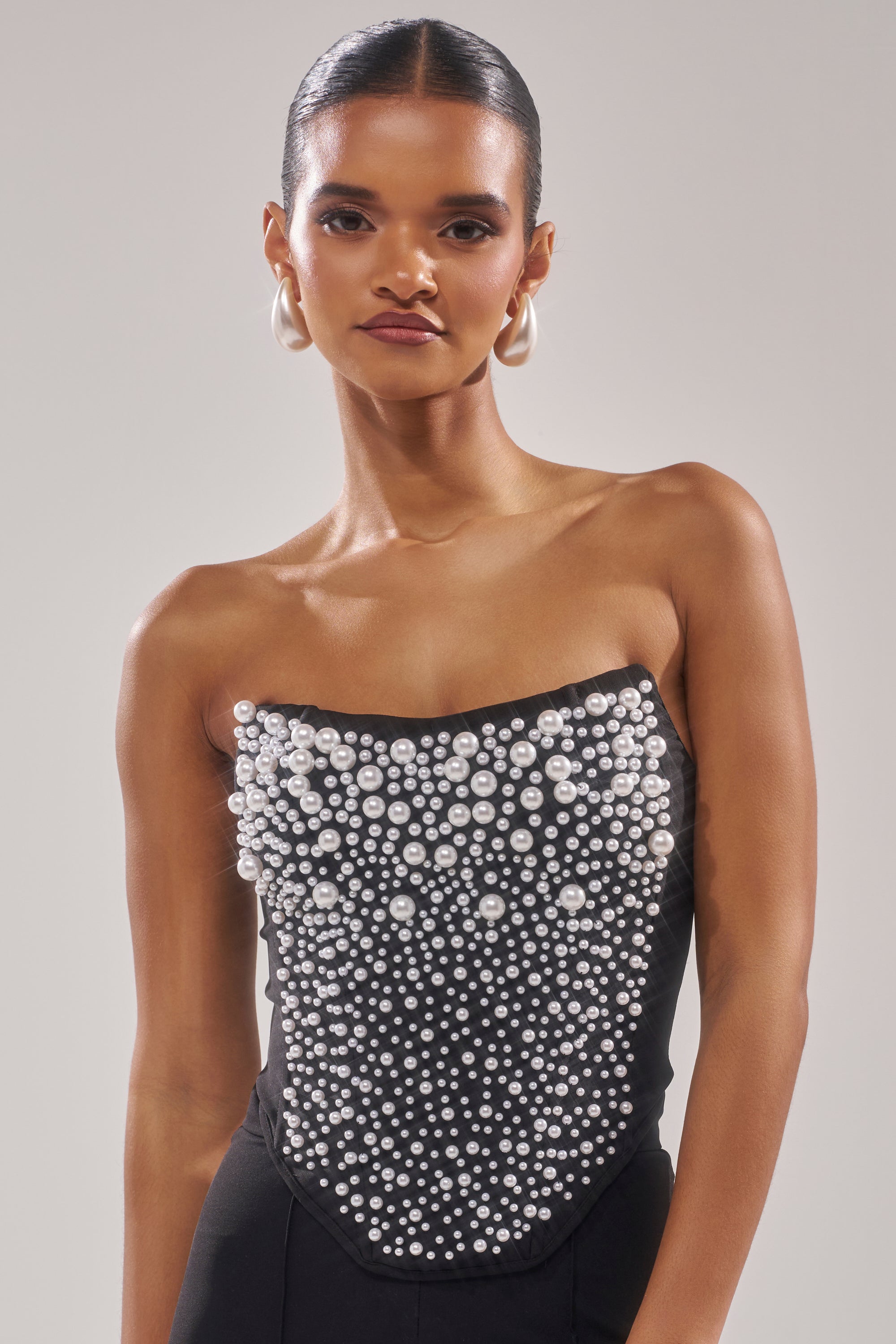 A woman with slicked-back hair wears large pearl earrings and the NEVER YOURS ALL OVER PEARL CORSET, a strapless black piece featuring clusters of white faux pearl embellishments, standing against a plain light grey background. 0