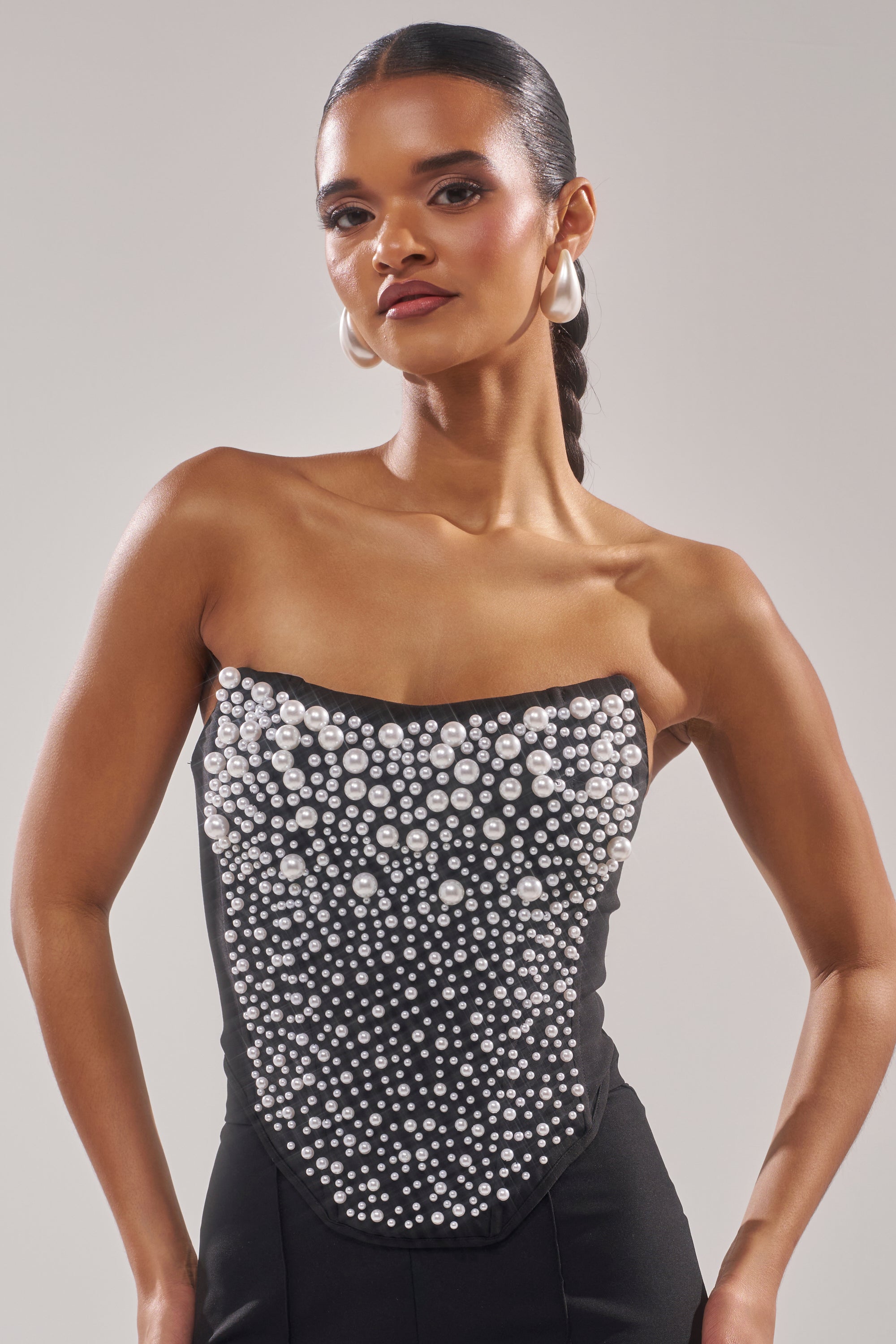A woman with slicked-back hair wears the NEVER YOURS ALL OVER PEARL CORSET, a strapless black piece adorned with clusters of faux pearls, pairing it with large hoop earrings as she poses confidently against a plain background. 1