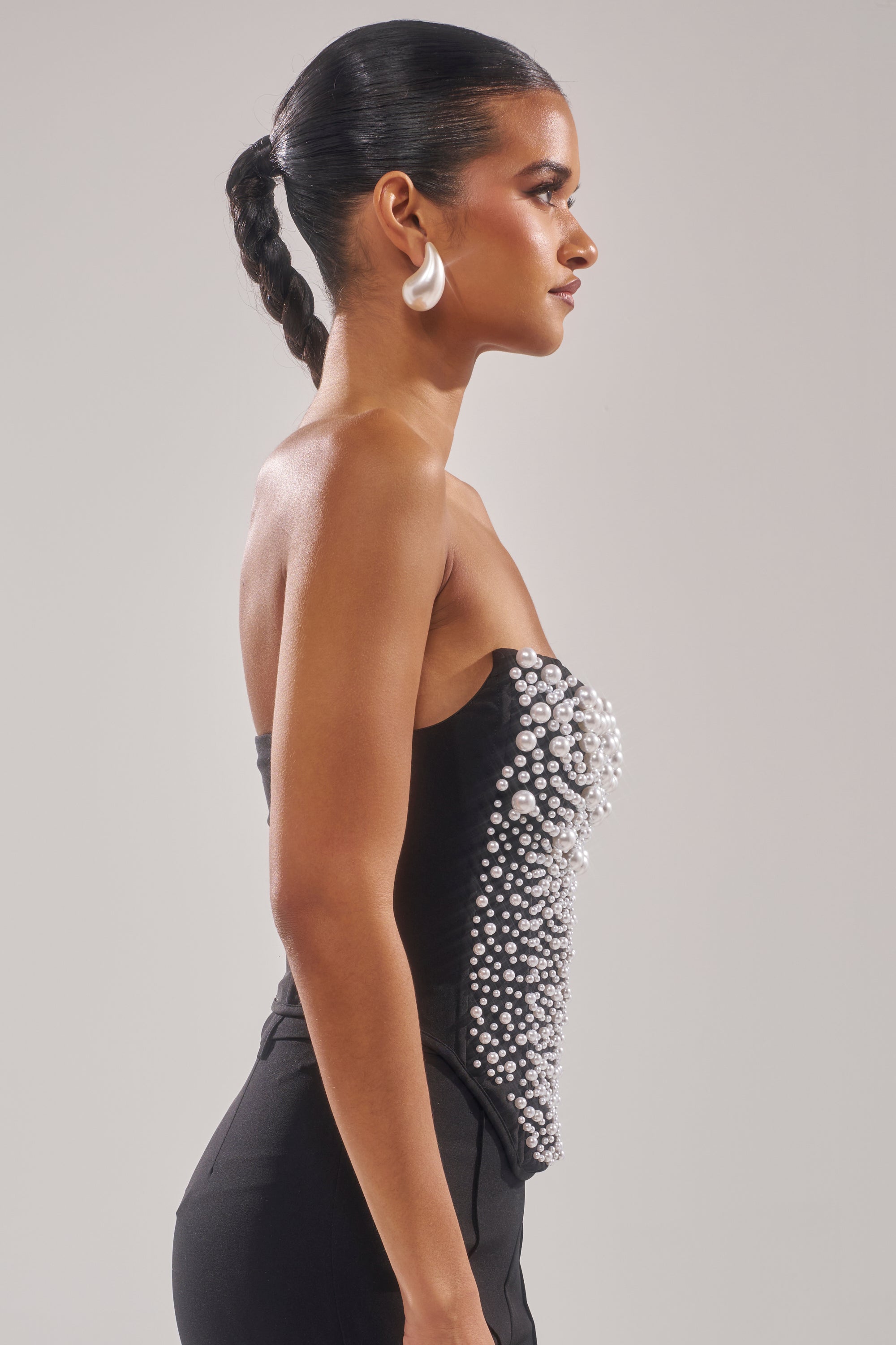 A woman with slicked-back braided hair stands in profile against a plain light background, wearing the NEVER YOURS ALL OVER PEARL CORSET with silver and faux pearl details, styled with large curved white earrings. 3
