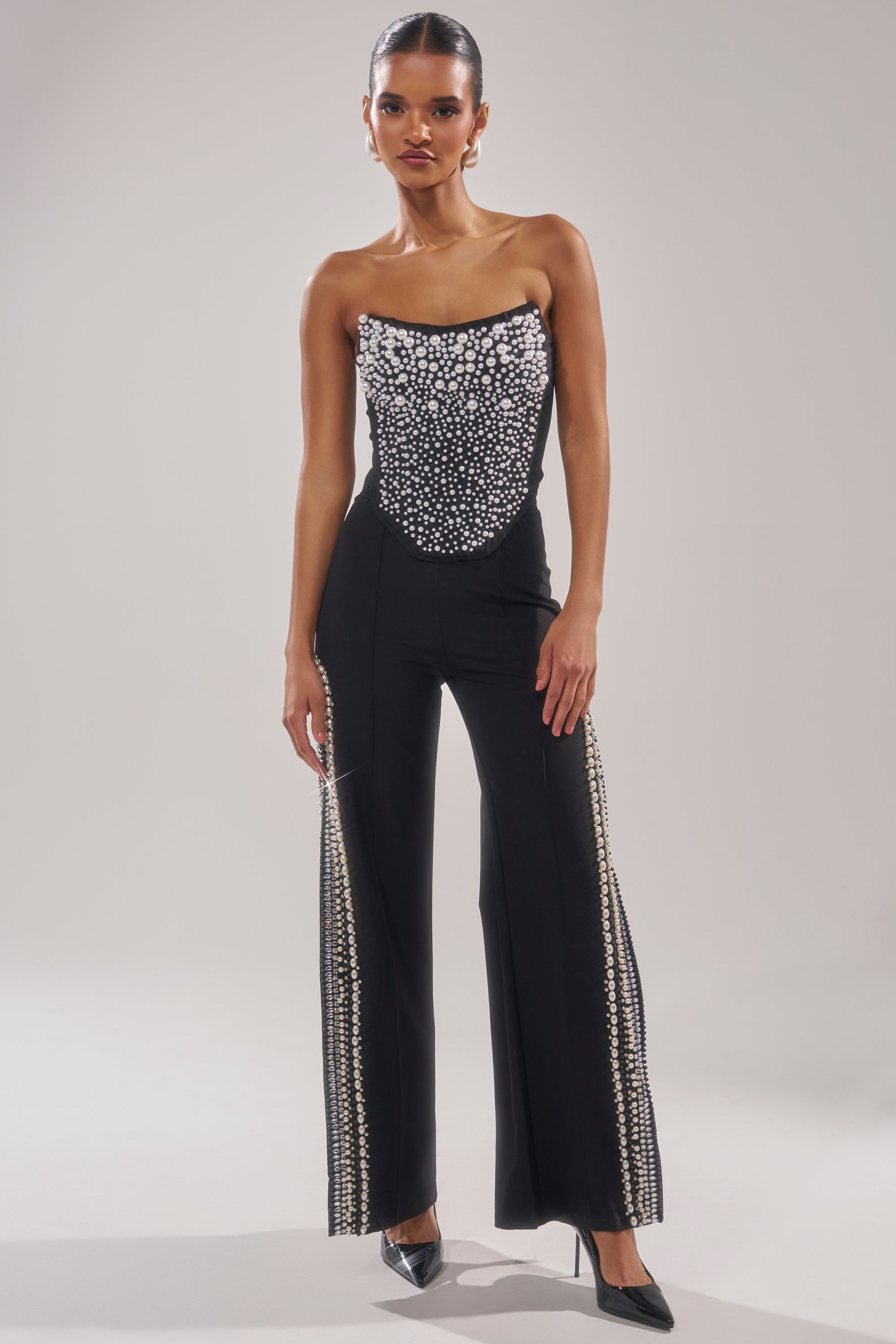 A woman poses against a plain light background wearing the NEVER YOURS ALL OVER PEARL CORSET, a strapless black jumpsuit with a fitted, beaded bodice, faux pearl embellishments, wide-leg pants with embellished panels, and black pointed heels. 5
