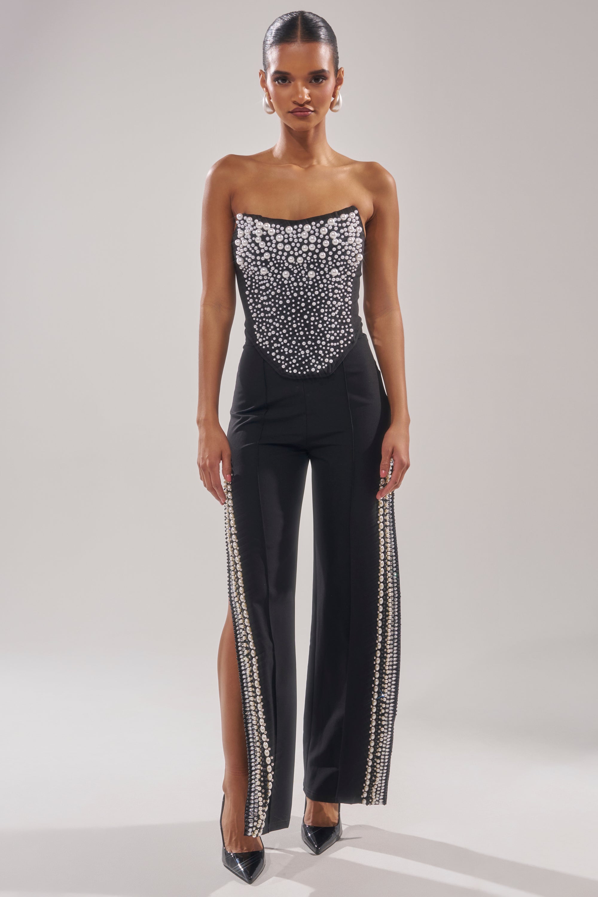 A woman faces forward in the NEVER YOURS ALL OVER PEARL CORSET, a strapless black jumpsuit with a faux pearl embellished bodice and vertical beads on front-slit pants. Her hair is sleek and pulled back, paired with black pointed heels. 6