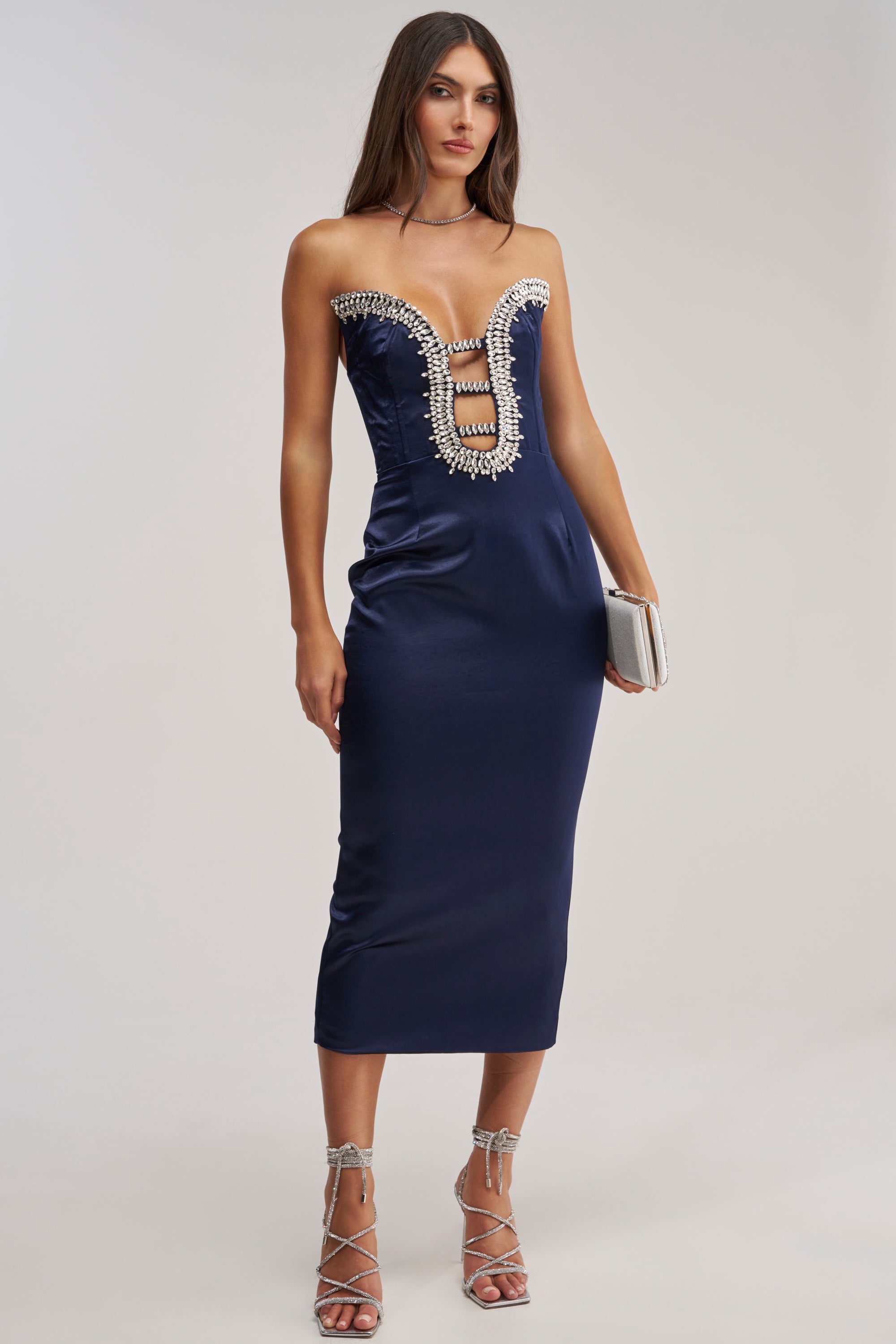 A woman wears the FEELIN THE LOVE RHINESTONE SATIN MIDI DRESS IN NAVY, featuring a strapless design, plunging neckline, crystal accents, and horizontal bands. She accessorizes with a silver clutch and strappy silver heels.