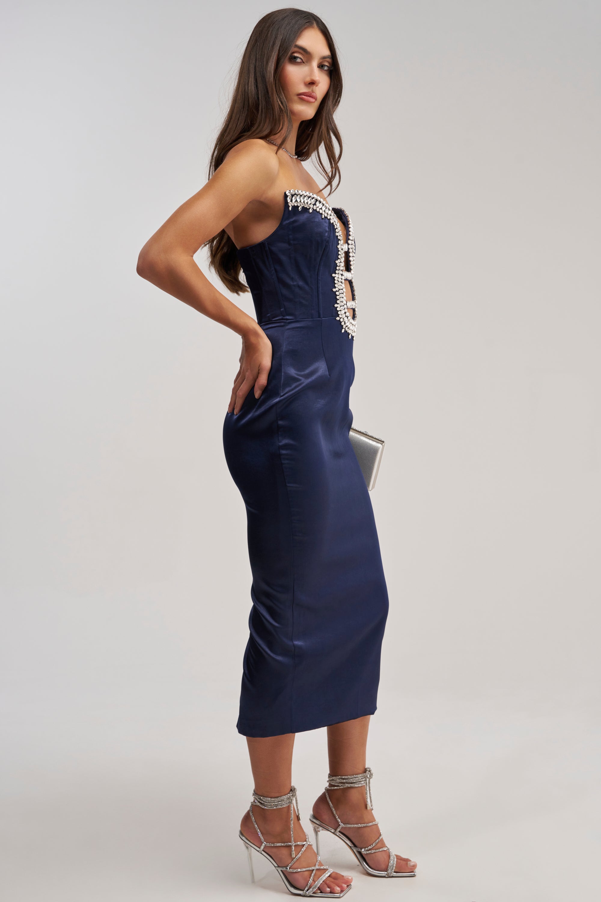 A woman poses in the FEELIN THE LOVE RHINESTONE SATIN MIDI DRESS IN NAVY, a fitted strapless navy midi dress with rhinestone details. She accessorizes with a small silver clutch and strappy silver heels against a light background.