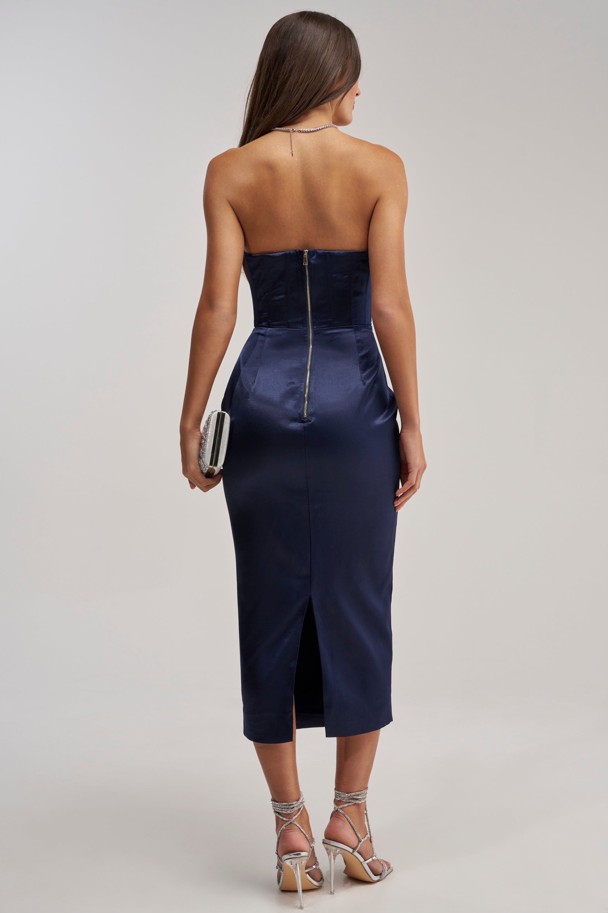 A woman stands facing away in the FEELIN THE LOVE RHINESTONE SATIN MIDI DRESS IN NAVY—a fitted, strapless satin midi with a back zipper and slit—paired with a silver clutch and strappy silver heels.