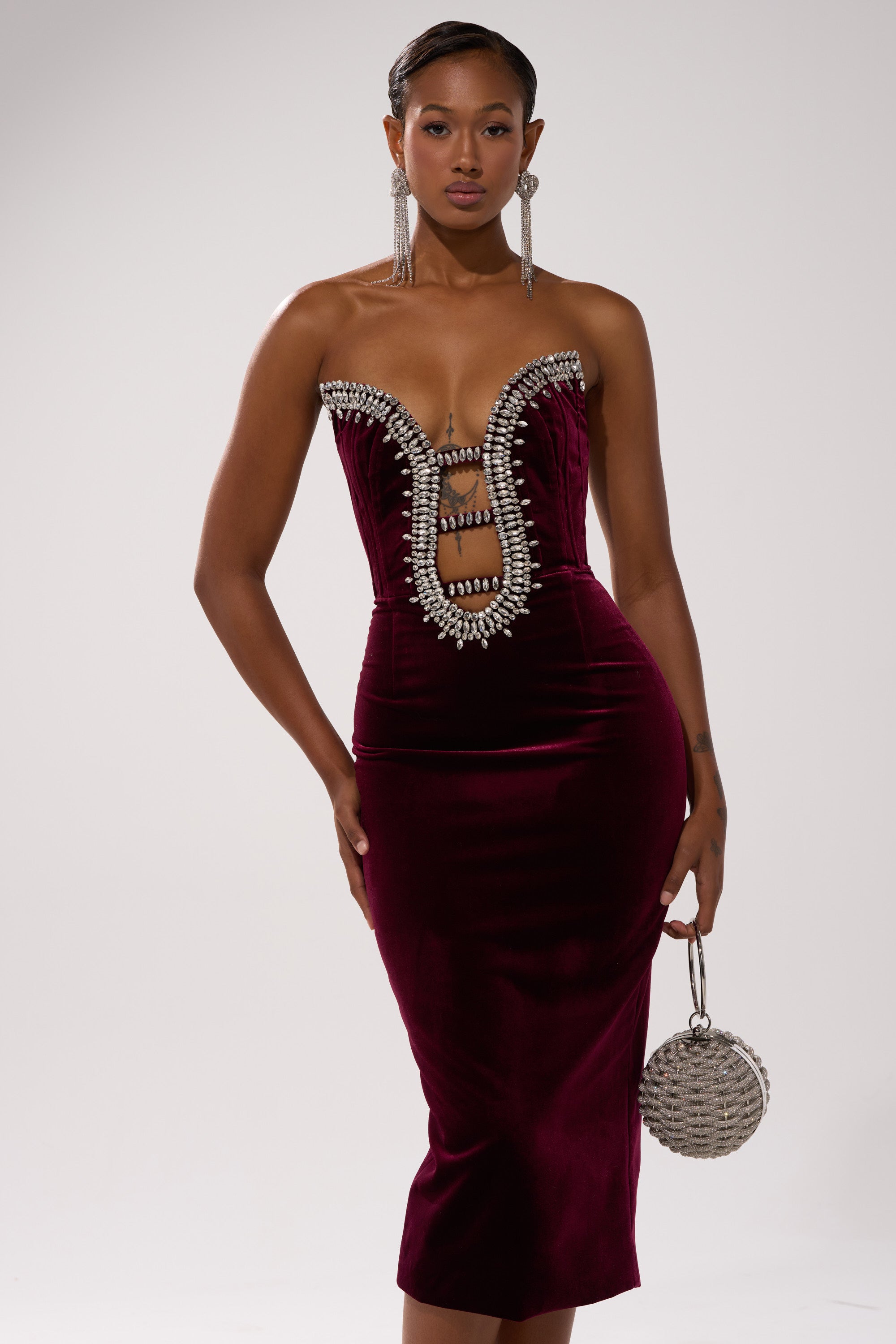 A woman stands confidently in the FEELIN THE LOVE VELVET RHINESTONE MINI DRESS—strapless, deep red, and embellished with silver rhinestones—accessorized with a round silver handbag and long, dangling earrings against a plain background.