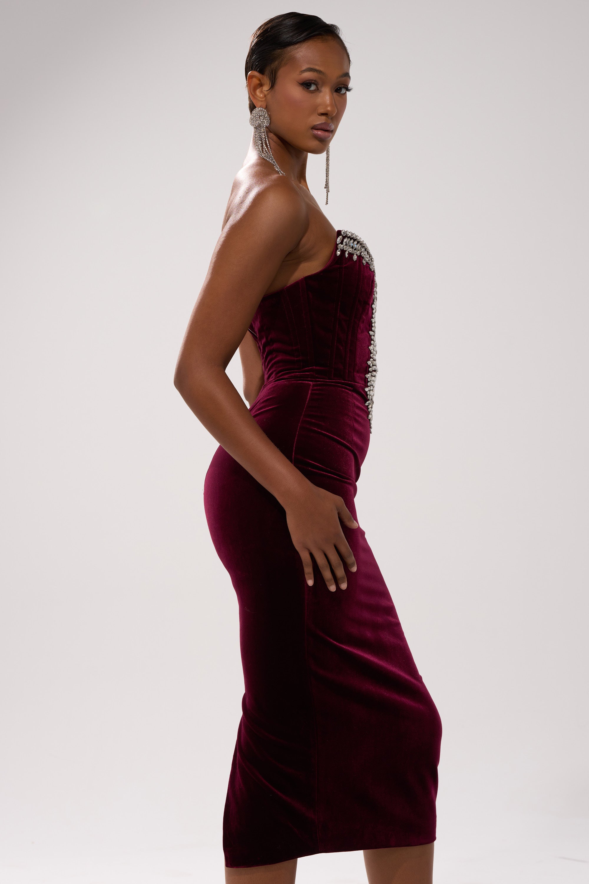 A woman models the FEELIN THE LOVE VELVET RHINESTONE MINI DRESS by AKIRA Label—a strapless burgundy velvet bodycon adorned with rhinestones—posing in profile against a light background, statement earrings on, exuding confidence.
