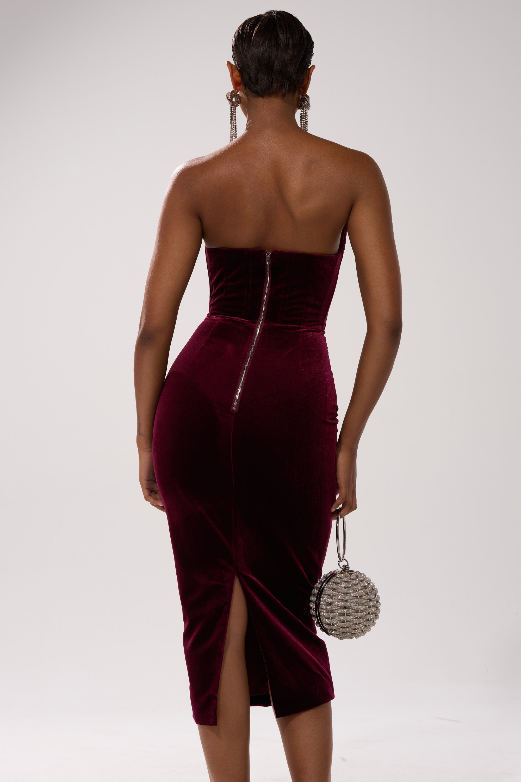 A woman is shown from behind wearing the FEELIN THE LOVE VELVET RHINESTONE MINI DRESS, a strapless deep burgundy velvet bodycon with a back zipper and hem slit. She carries a silver studded handbag and wears long dangling earrings.