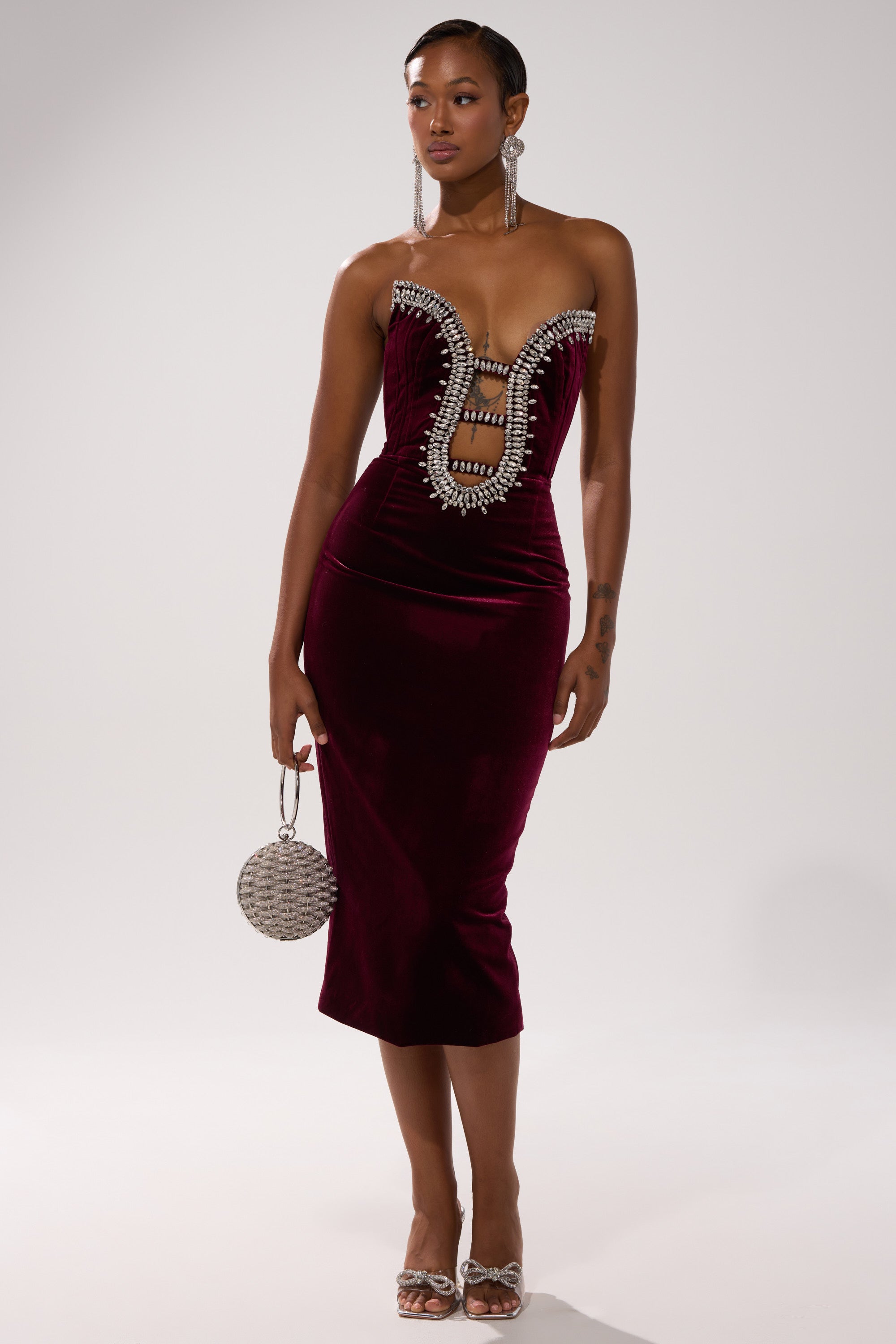 Wearing the FEELIN THE LOVE VELVET RHINESTONE MINI DRESS, a woman with statement earrings, open-toe heels, and a round silver handbag poses against a plain light background.