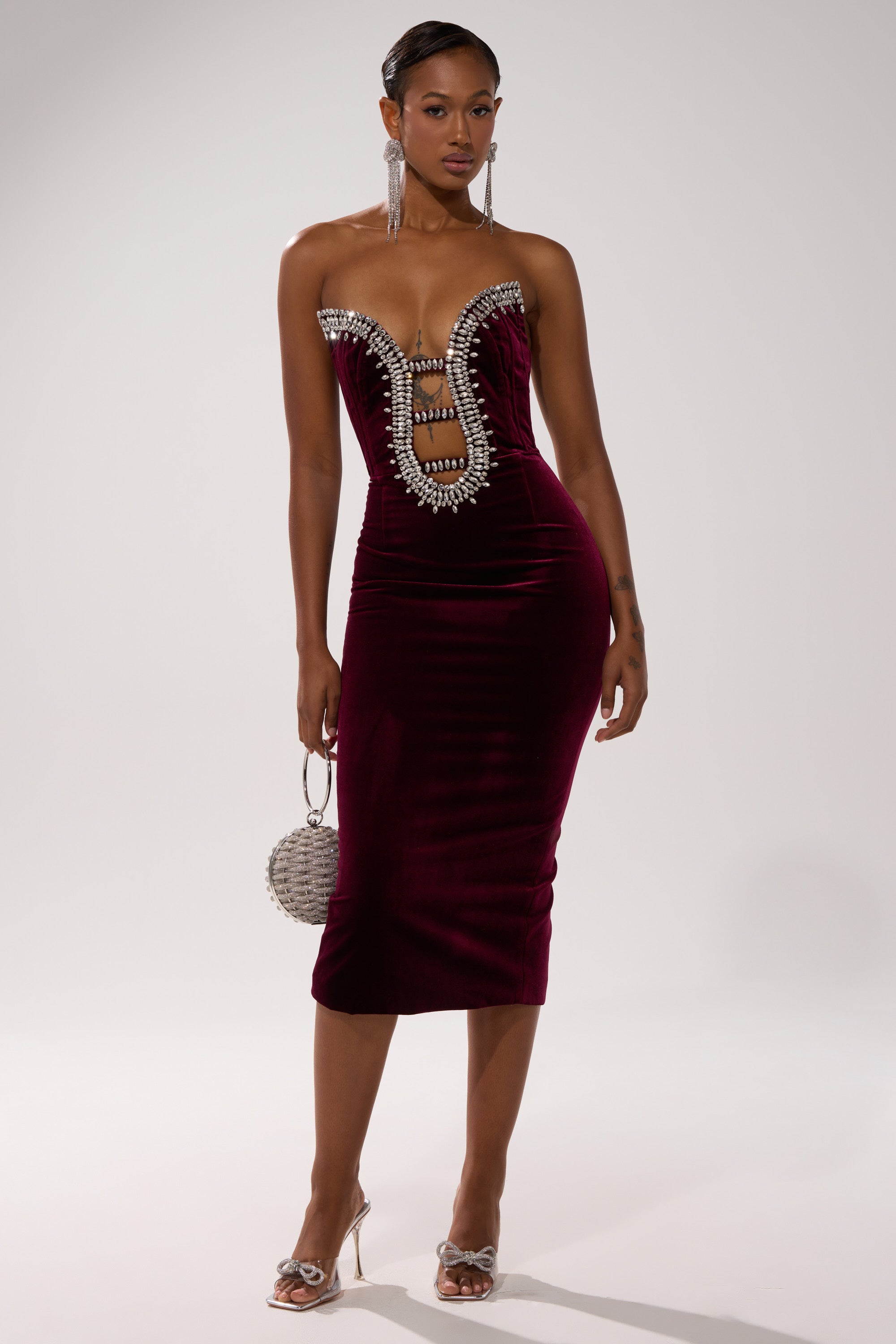 A woman models the FEELIN THE LOVE VELVET RHINESTONE MINI DRESS, a strapless deep red velvet bodycon with bold ladder cutouts and sparkling rhinestones. She pairs it with a round beaded handbag, silver heels, and long earrings against a plain white background.