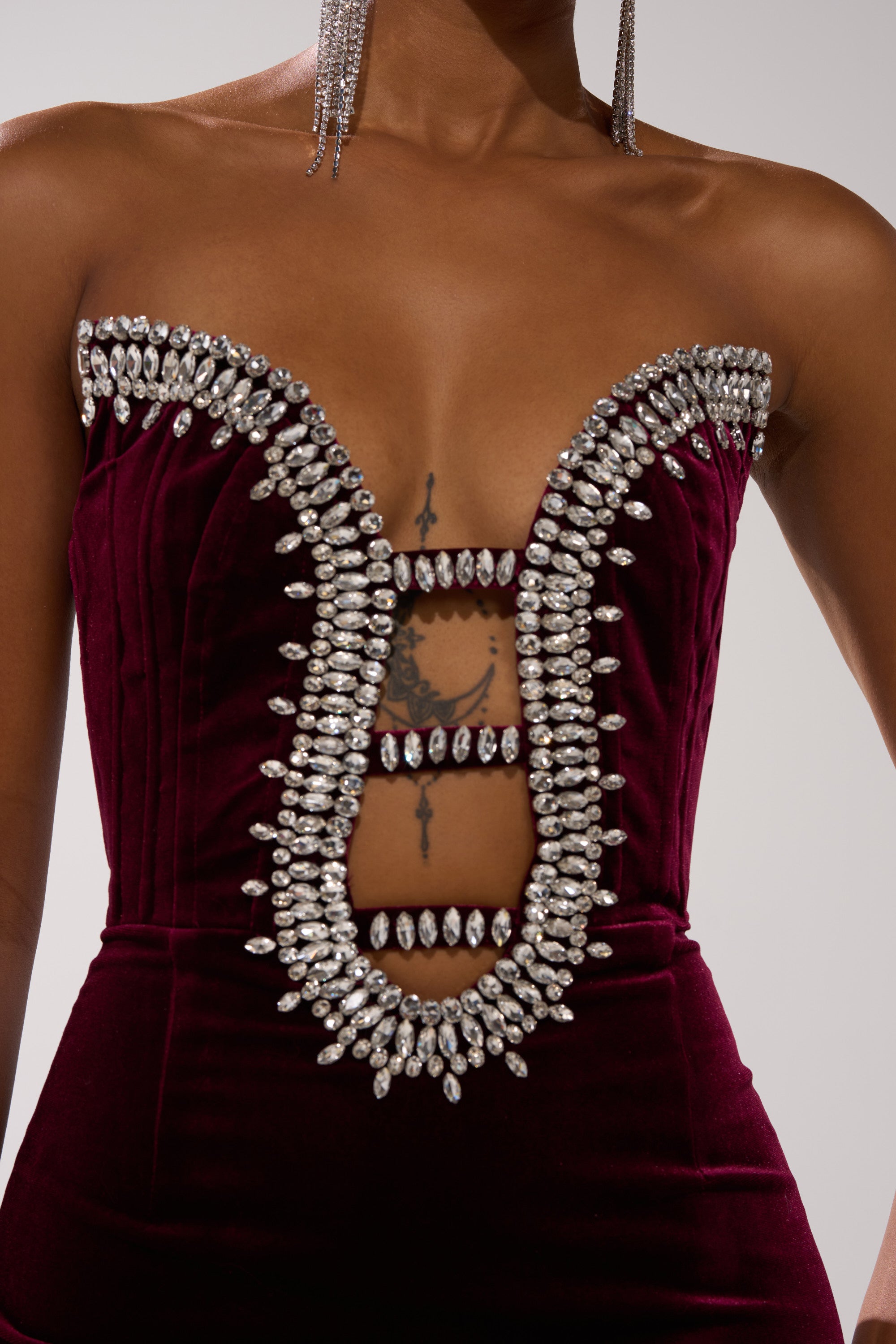 Wearing the FEELIN THE LOVE VELVET RHINESTONE MINI DRESS—a strapless, deep burgundy mini with a rhinestone-trimmed plunging neckline that reveals a chest tattoo—paired with long rhinestone earrings for a bold, glamorous look.