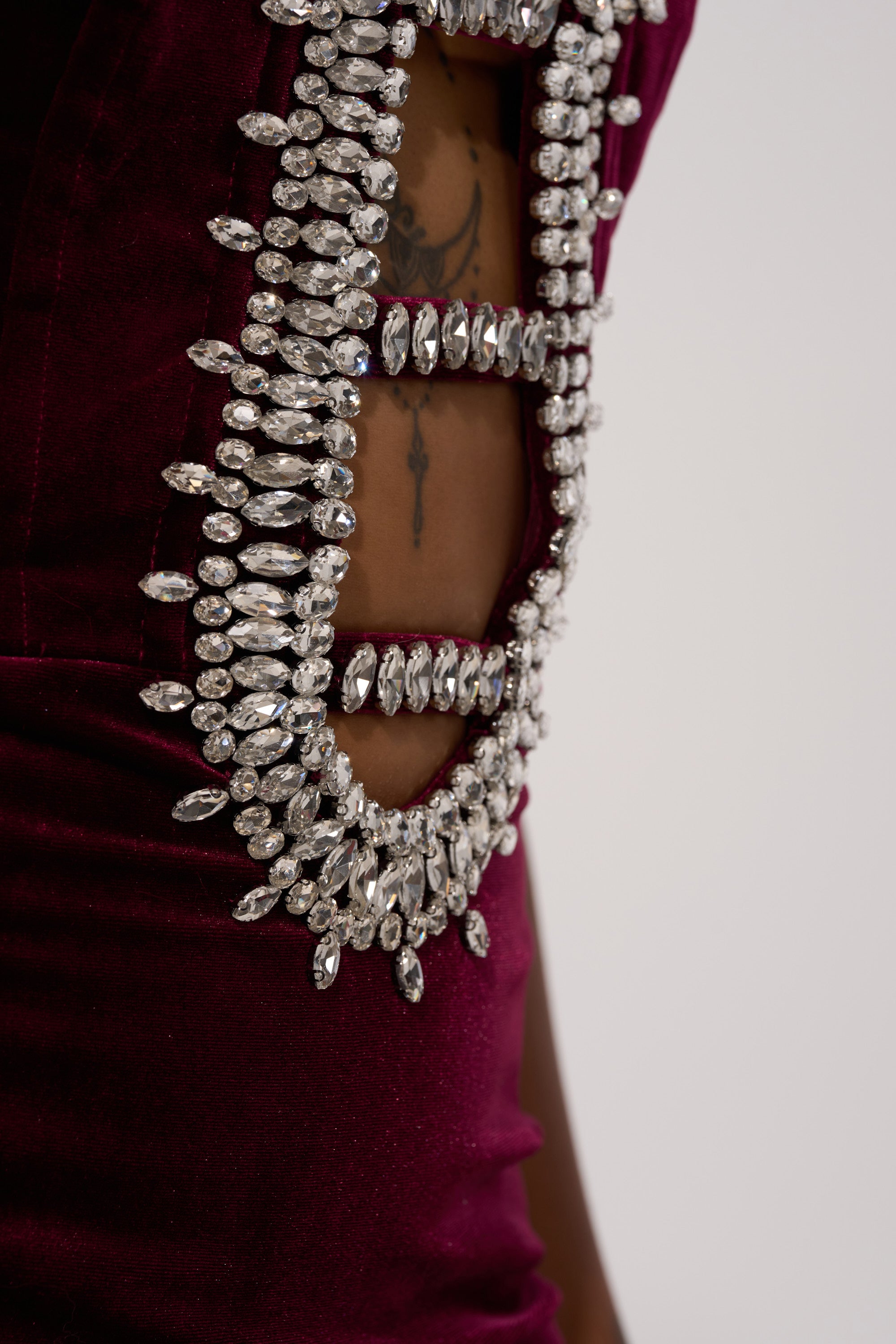 A close-up shows a person wearing the FEELIN THE LOVE VELVET RHINESTONE MINI DRESS in deep burgundy with oval crystal embellishments framing a cutout, revealing a side tattoo. The plain, neutral background highlights the glamorous look.