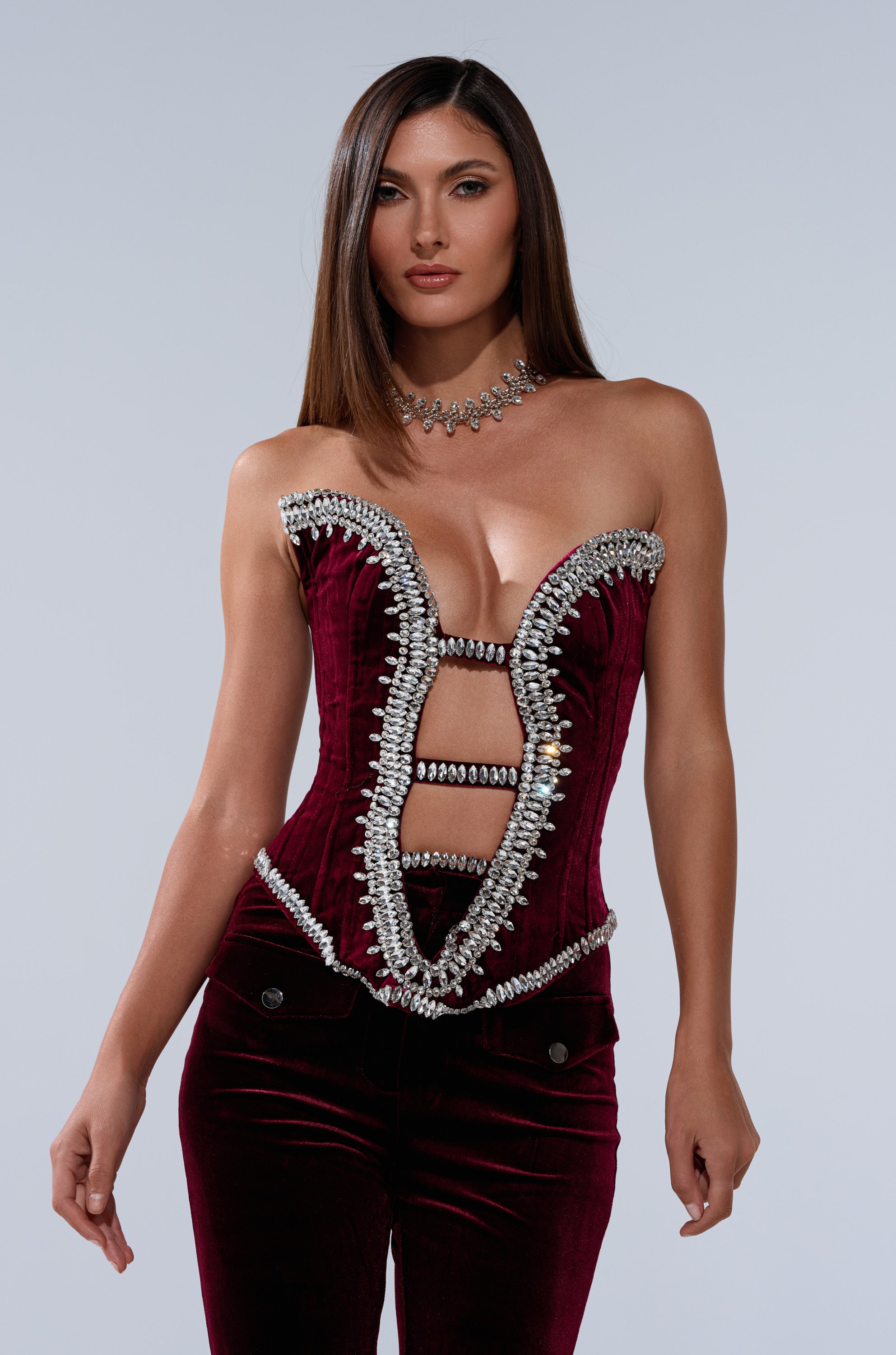 A woman with long, straight brown hair wears the LUXE LIFE CORSET IN BURGUNDY, a crystal-embellished velvet corset, paired with matching velvet pants. She stands against a plain, light gray background.