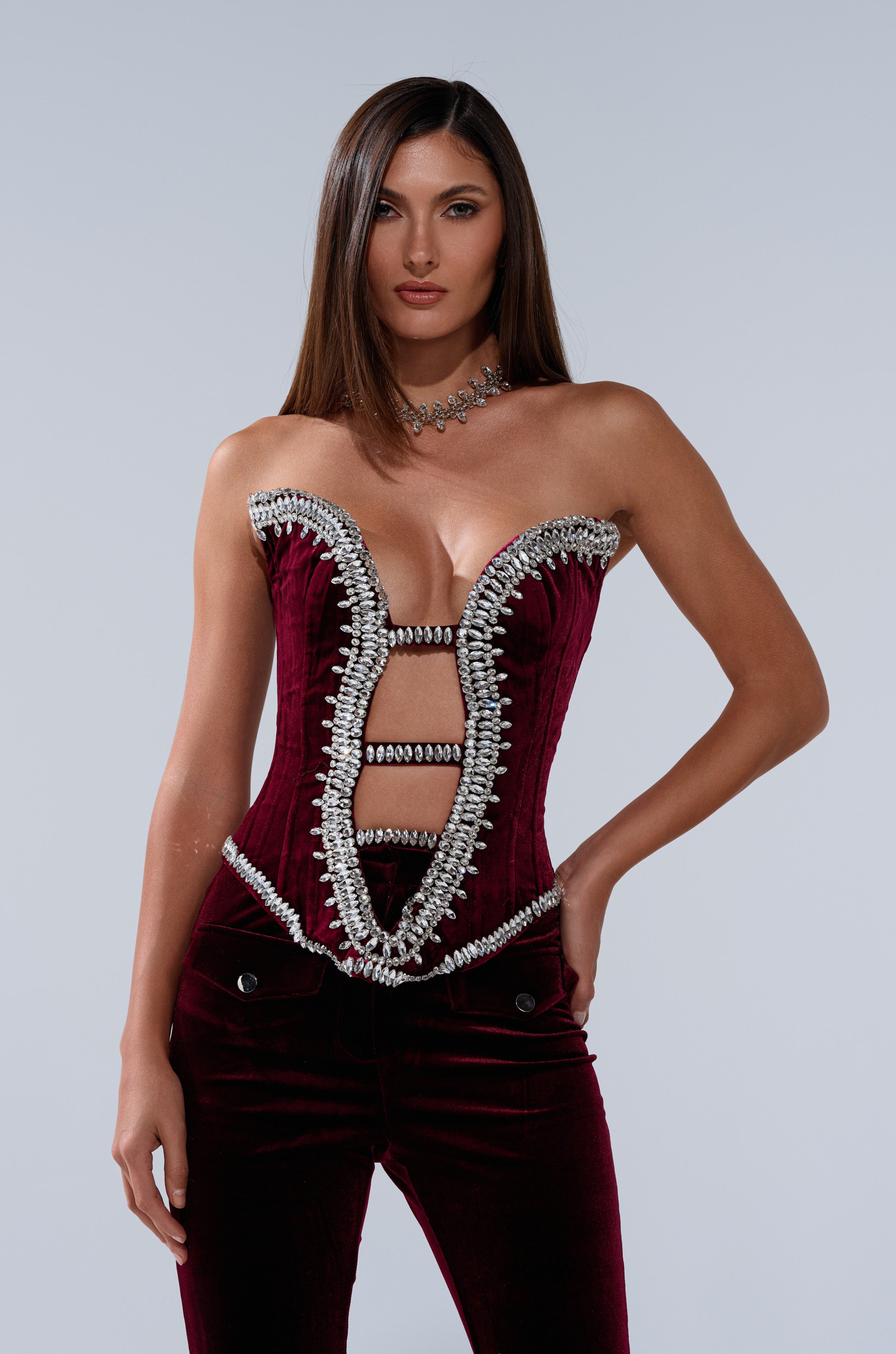 A woman with straight brown hair models the LUXE LIFE CORSET IN BURGUNDY, featuring silver detailing and a unique cutout design, paired with matching pants and a silver necklace against a plain light background.