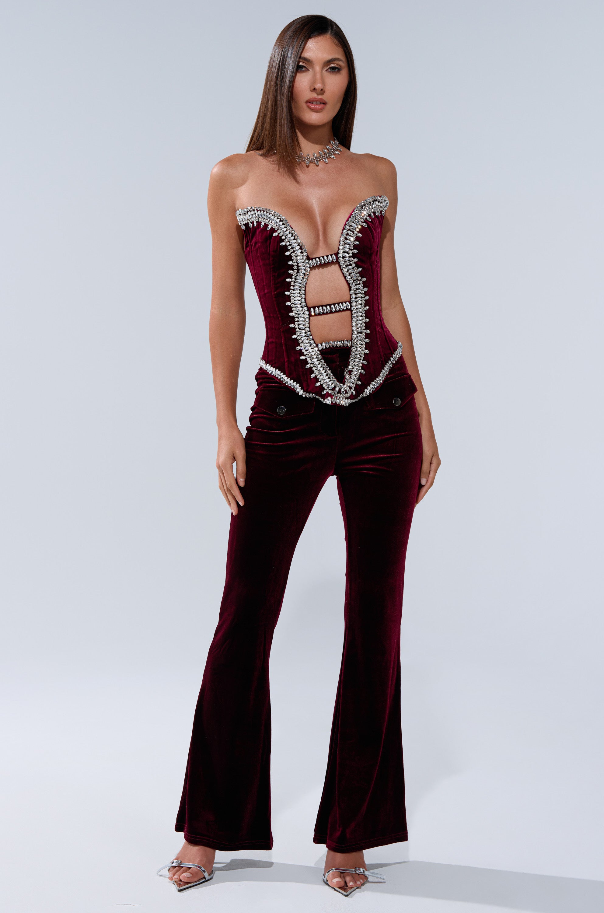 A woman stands wearing the LUXE LIFE CORSET IN BURGUNDY with a plunging, structured neckline, crystal accents, and flared pants. Her straight, shoulder-length hair and high-heeled sandals complete the look.