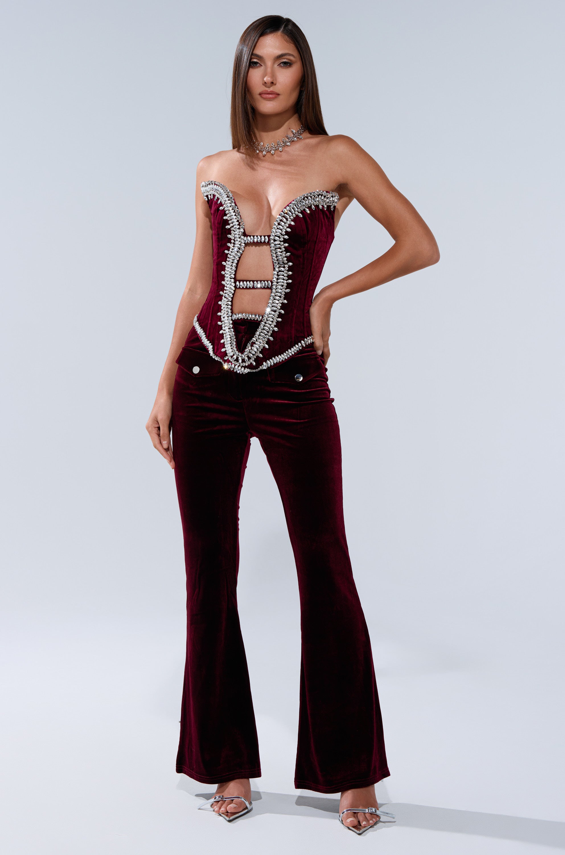 A woman models the LUXE LIFE CORSET IN BURGUNDY—a velvet jumpsuit with a plunging, crystal-embellished neckline and flared pants—paired with silver heels against a plain light background.