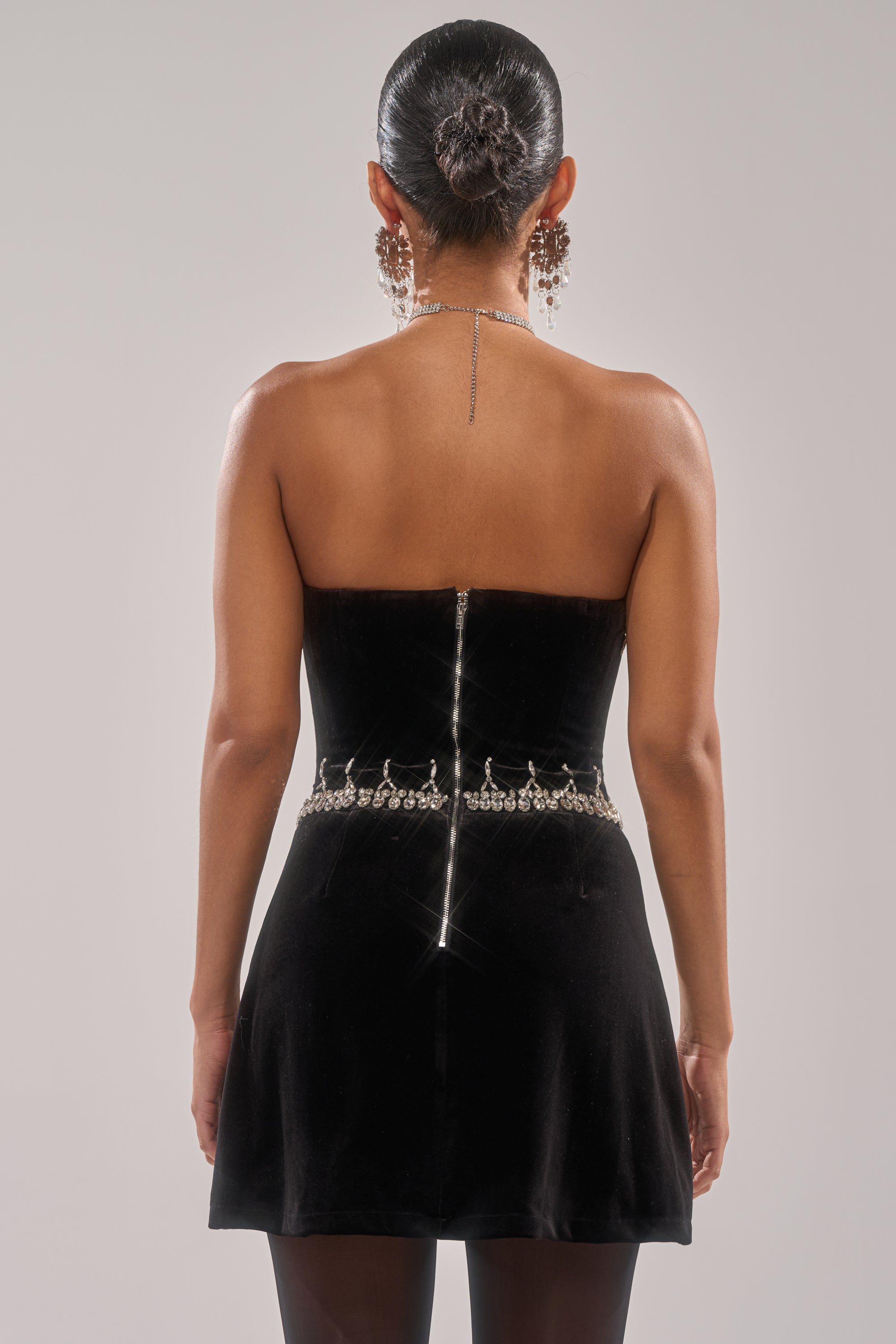 A woman faces away, wearing the HIGH SOCIETY VELVET RHINESTONE MINI DRESS with a jeweled belt, earrings, and a long necklace draped down her back. Her hair is in a sleek bun—an elegant look for any evening event. 3
