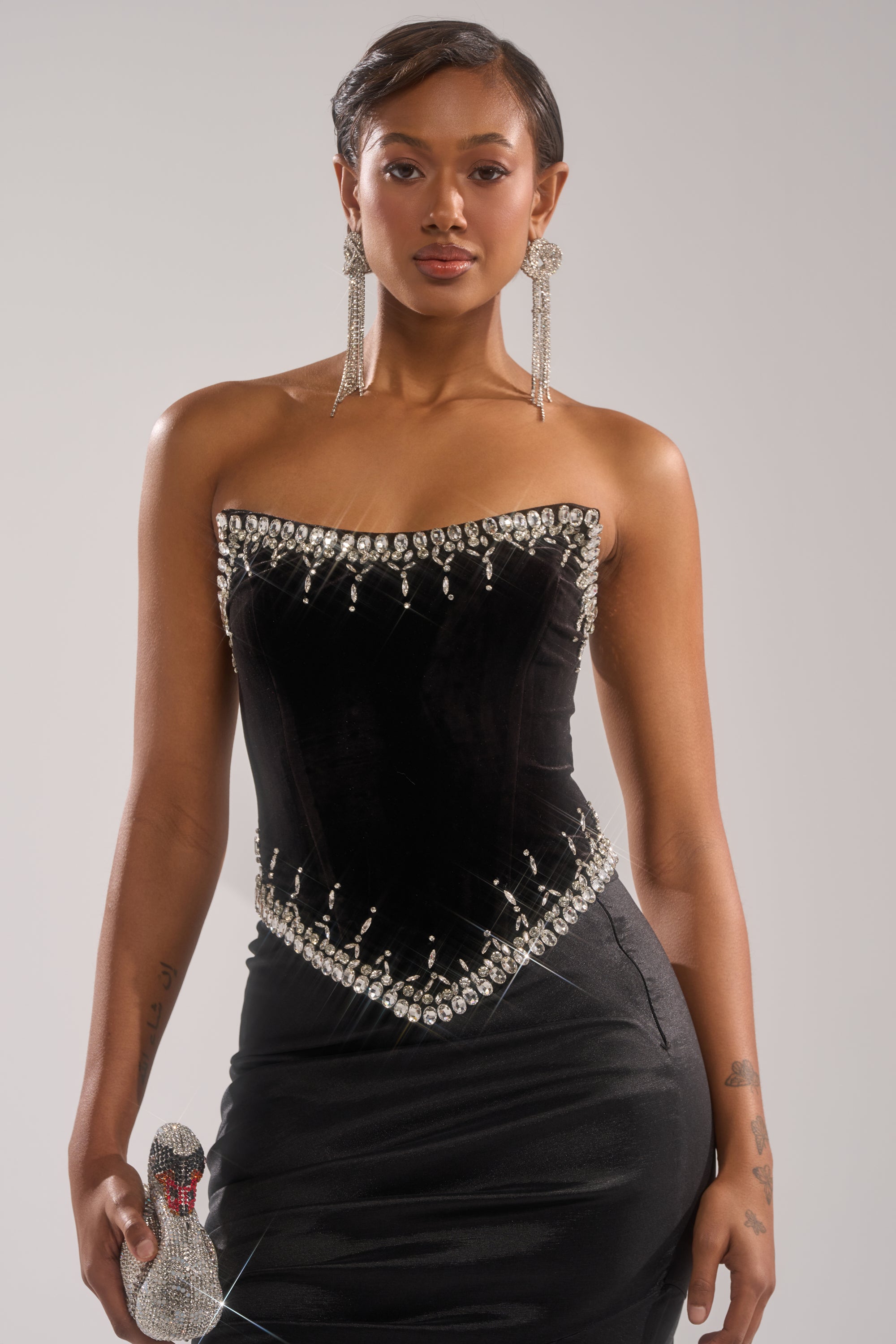 A woman wears the LUX LIFE CORSET IN BLACK, a strapless velvet dress with rhinestone accents at the neckline and waist. She has long dangling earrings and holds a glittery high heel, set against a plain light background. 0