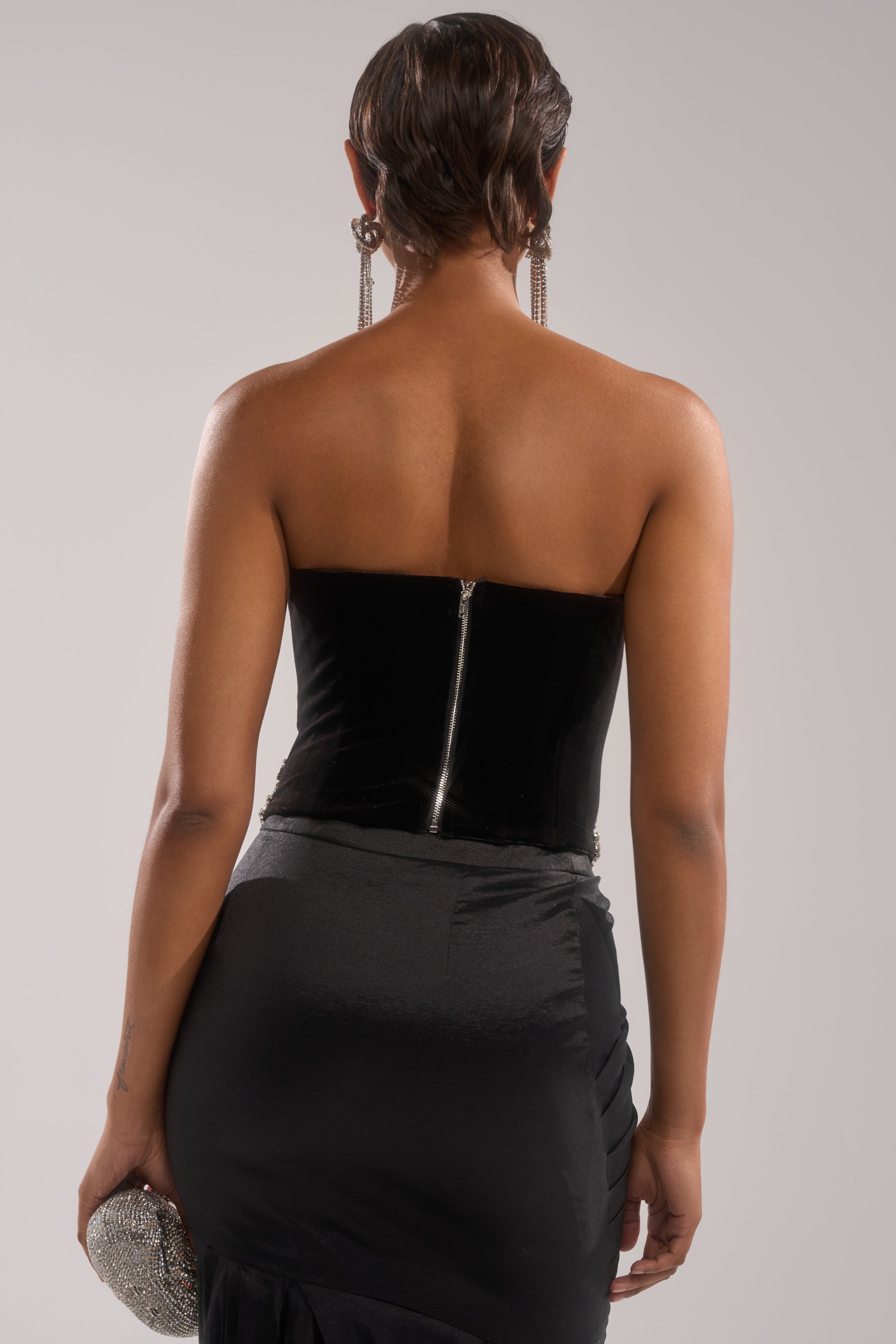 A person with short hair is seen from the back wearing the LUX LIFE CORSET IN BLACK, a strapless corset dress with a zipper, paired with long earrings and holding a sparkling clutch against a plain light background. 3
