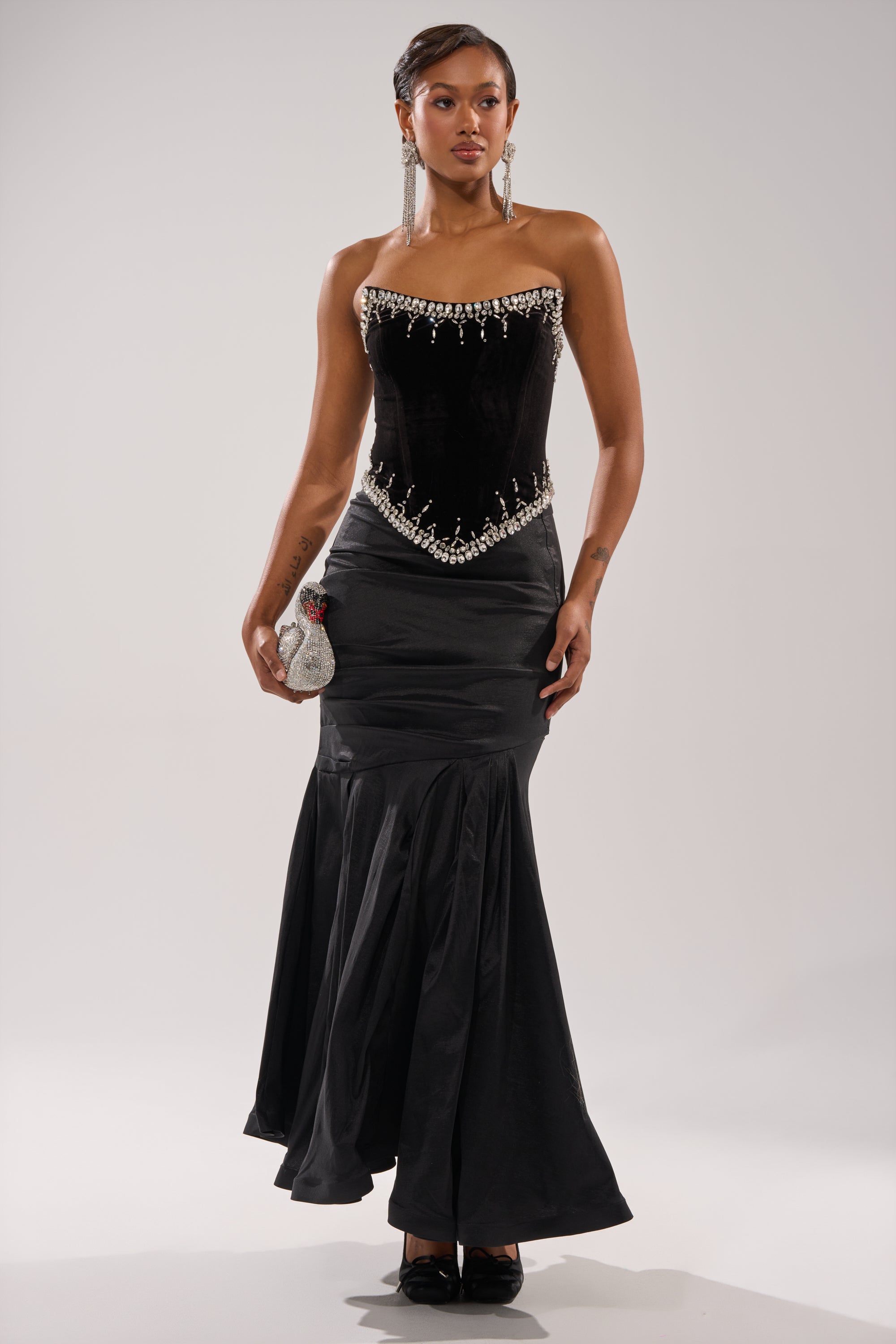 A woman poses against a plain background in the LUX LIFE CORSET IN BLACK, a strapless velvet gown with pearl accents and a flared satin skirt, accessorized with a swan-shaped handbag and long dangling earrings. 4