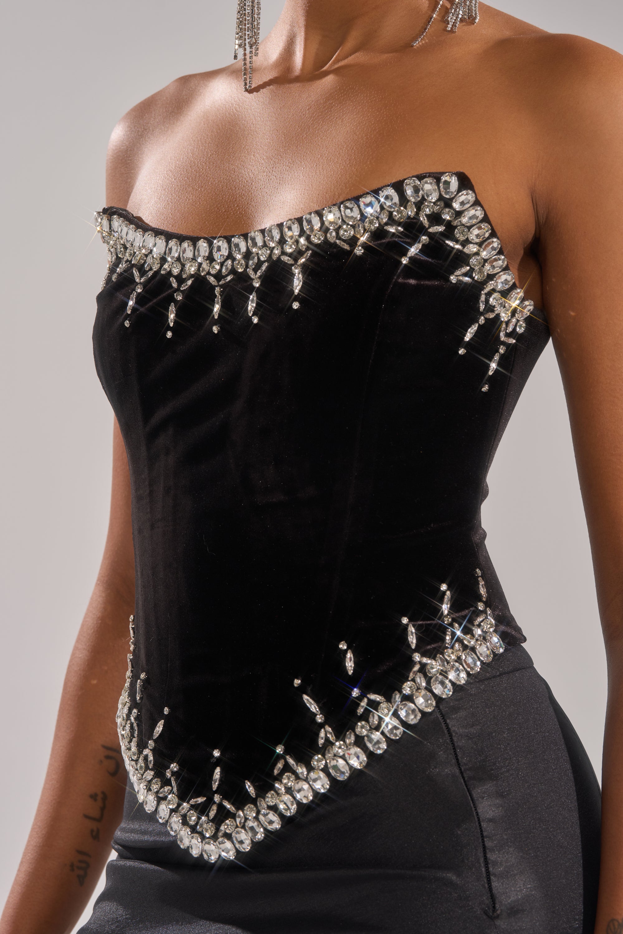 A woman wears the LUX LIFE CORSET IN BLACK—strapless and velvet, with sparkling rhinestones along the edges. She pairs it with a dark skirt and dangling earrings; part of a tattoo is visible on her arm. 5