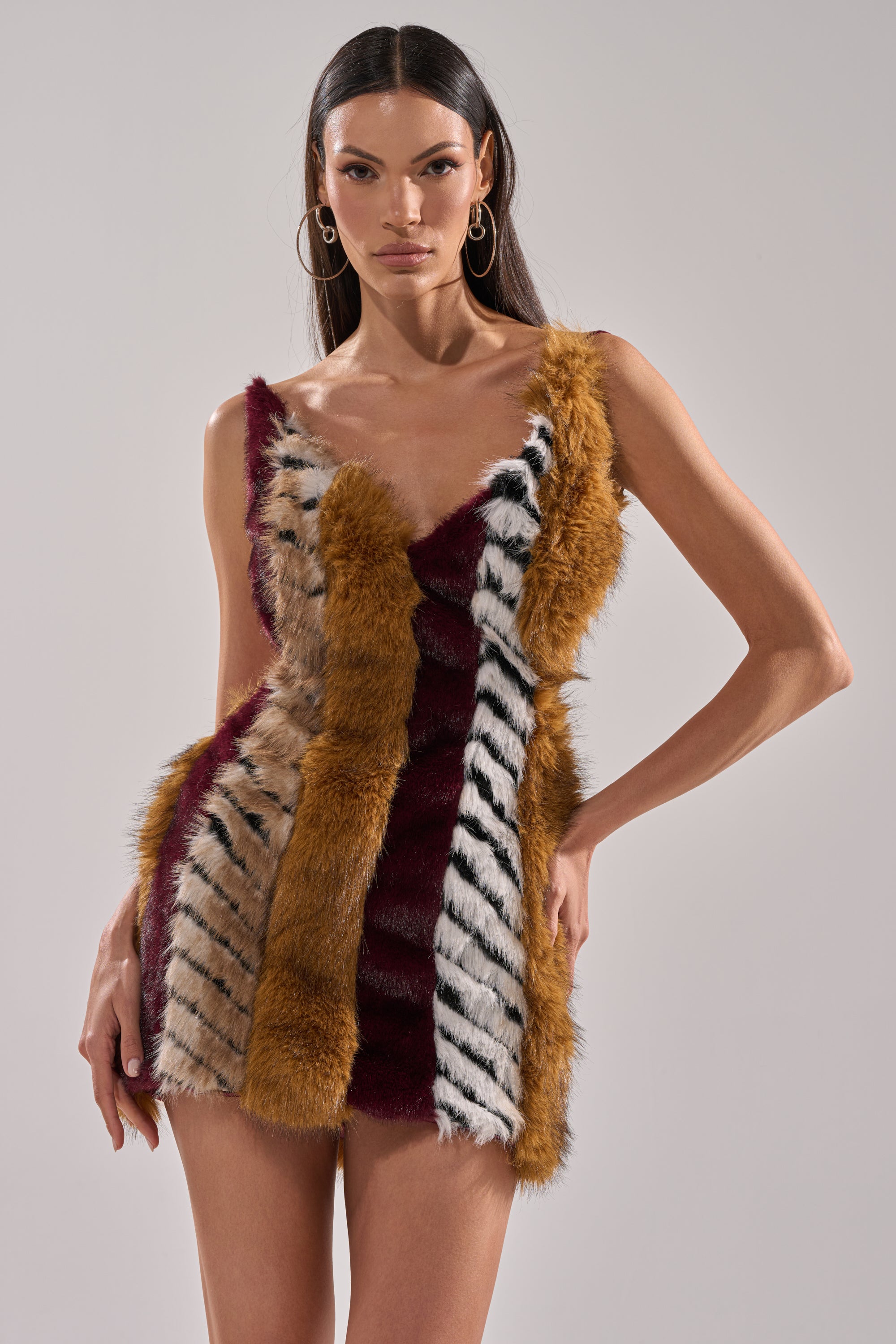 A woman stands confidently in the ANIMALISTIC MINI DRESS, a sleeveless satin mini with a deep neckline and vertical stripes of tan, burgundy, and black-and-white animal print faux fur.