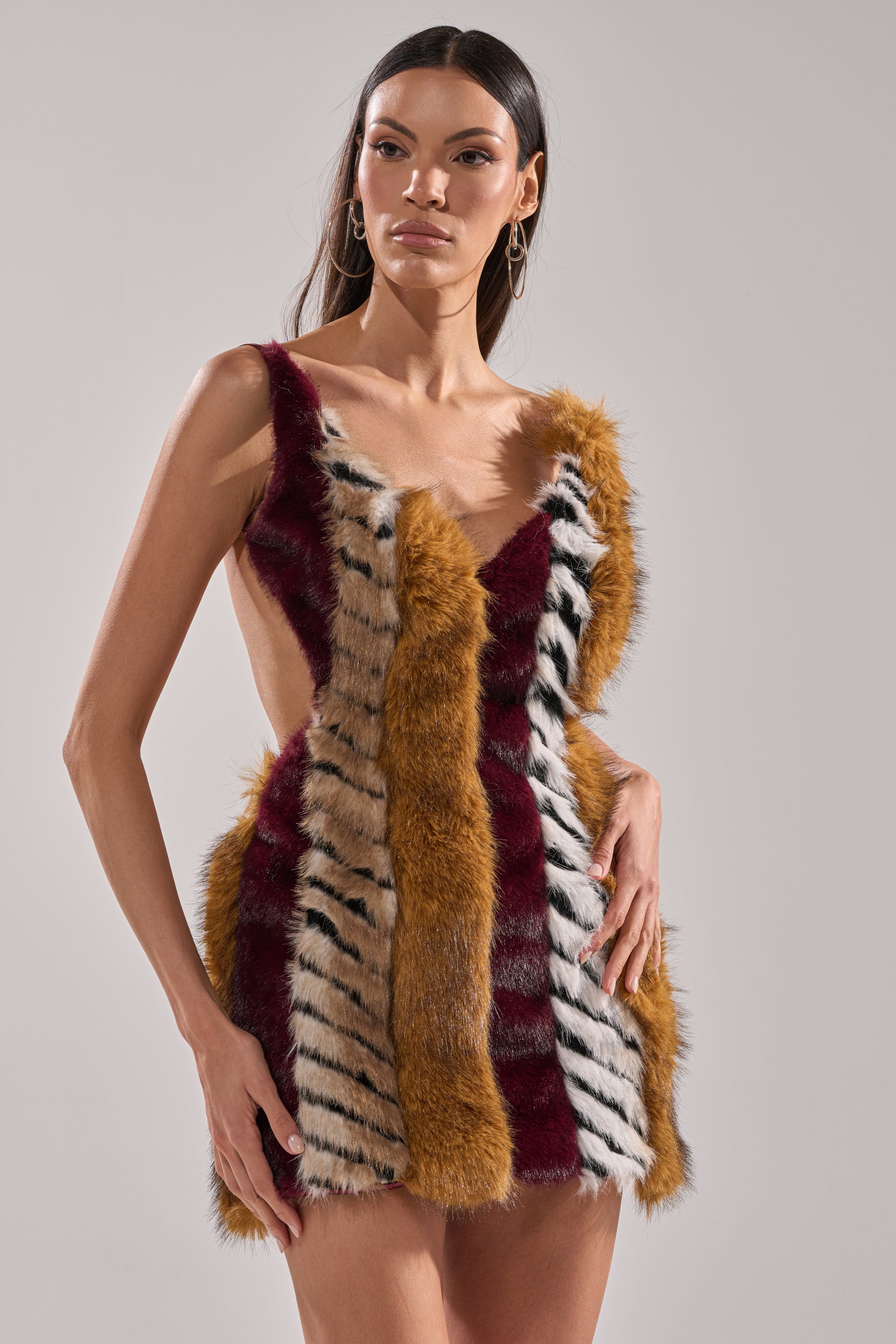A woman wears the ANIMALISTIC MINI DRESS, a sleeveless faux fur mini with vertical burgundy, tan, and white stripes with black accents, standing before a plain light background.