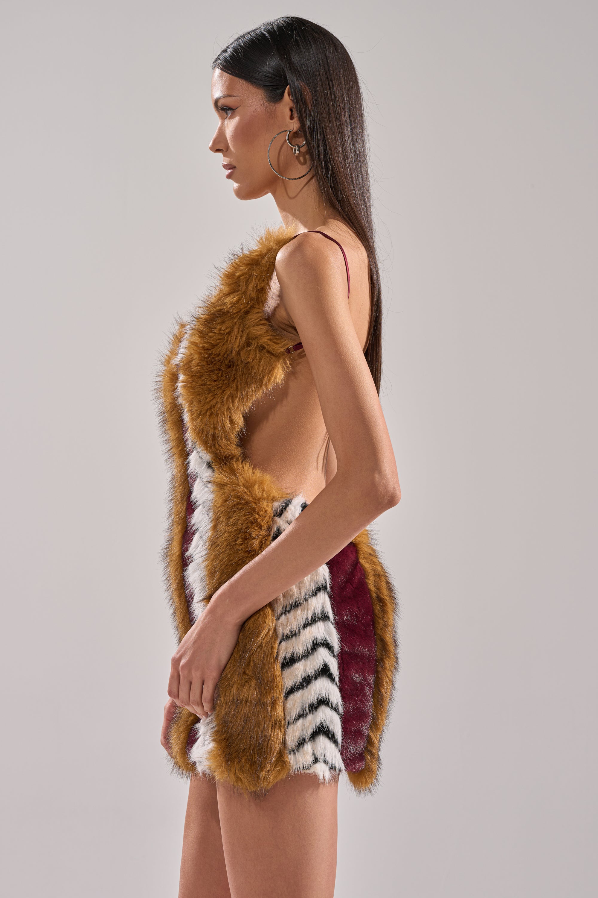 A woman with long dark hair and hoop earrings stands in profile against a plain light background, wearing the ANIMALISTIC MINI DRESS—a fitted mini dress made of multicolored faux fur in brown, white, black, and burgundy sections.