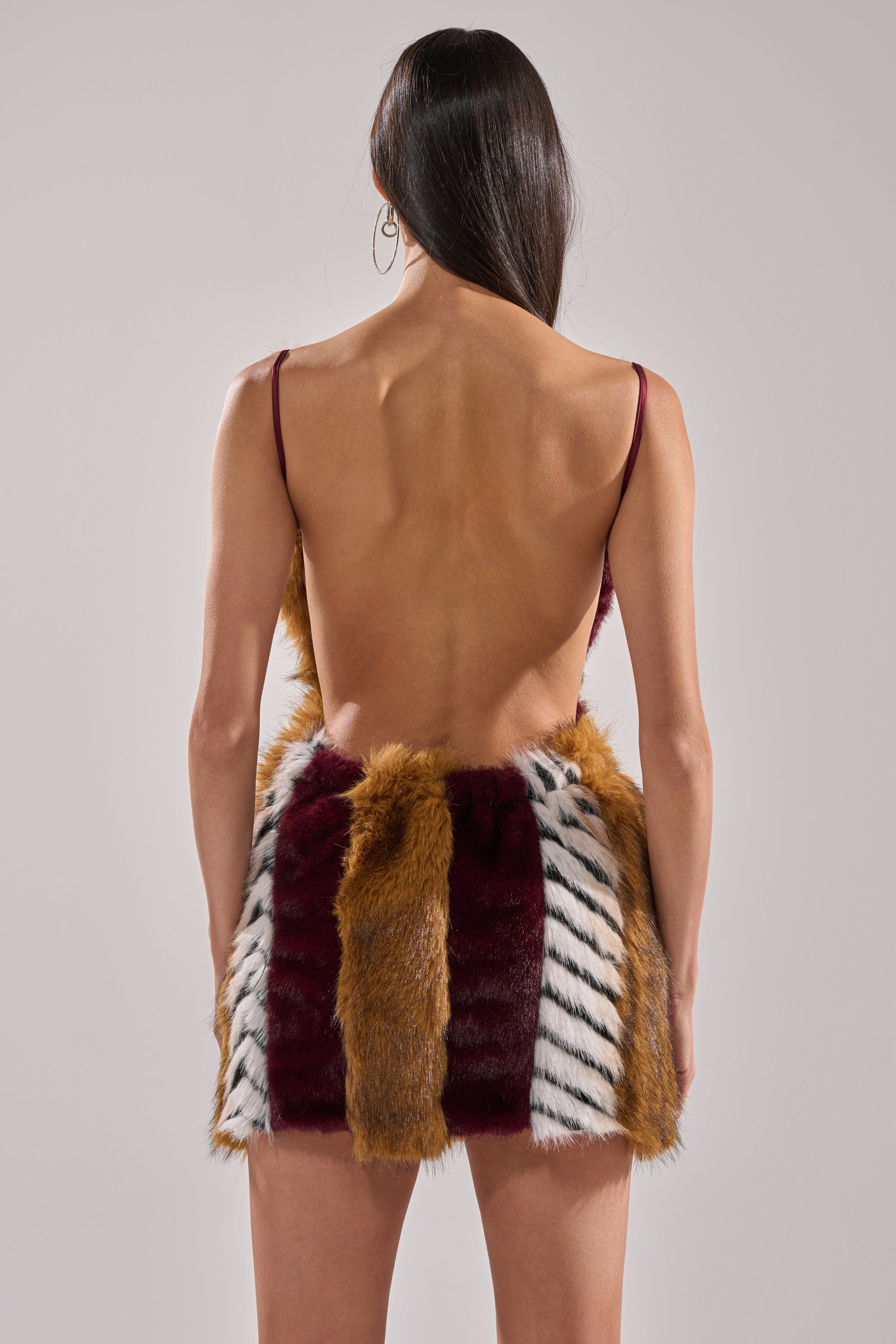 A woman with long dark hair faces away, wearing the ANIMALISTIC MINI DRESS—a form-fitting style with thin straps and panels of maroon, mustard yellow, and white faux fur featuring black stripes. The background is neutral.