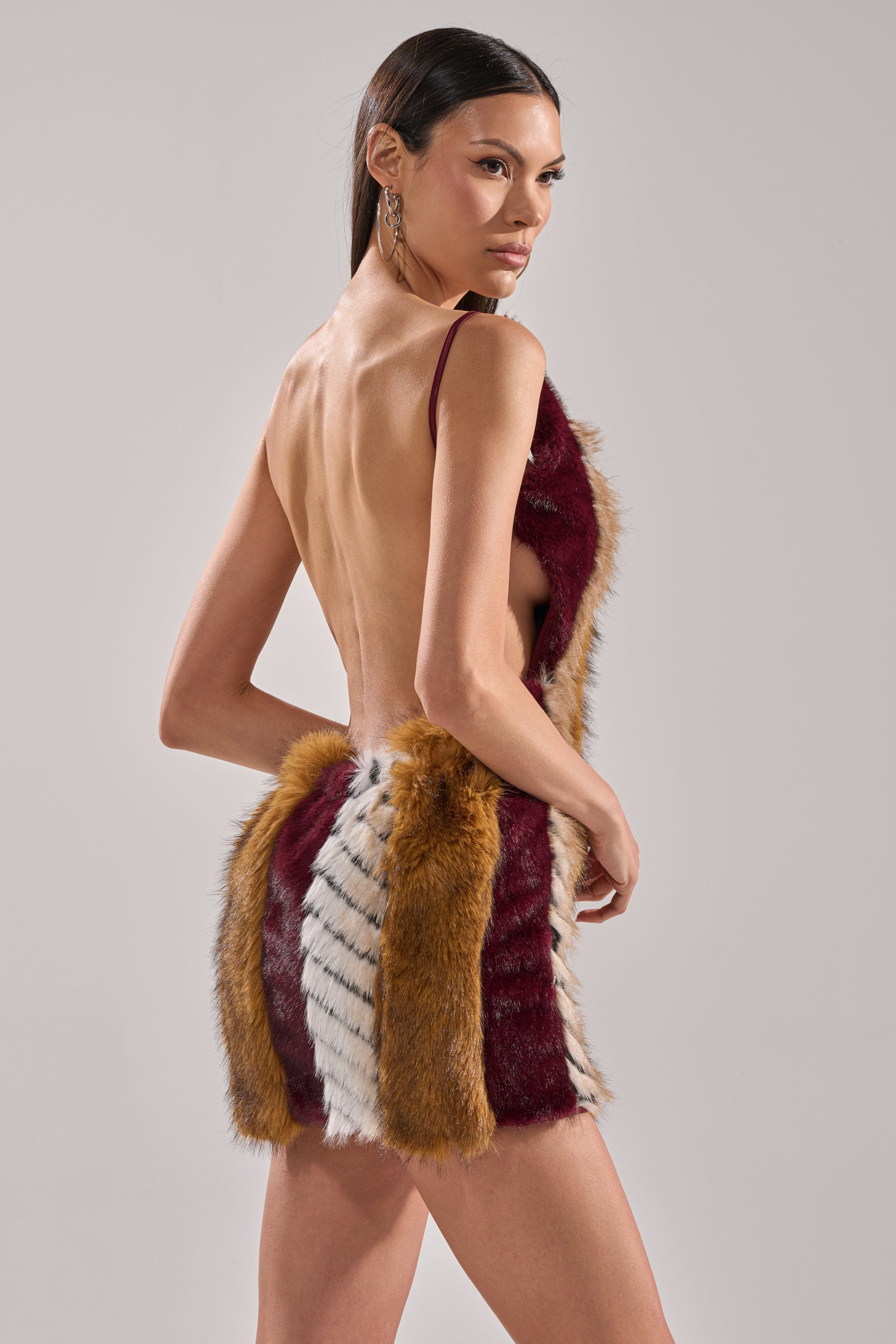 A woman models the ANIMALISTIC MINI DRESS, a sleeveless, backless mini with maroon, mustard, and white faux fur panels. She stands against a plain background, highlighting its body-hugging fit as she looks over her shoulder.