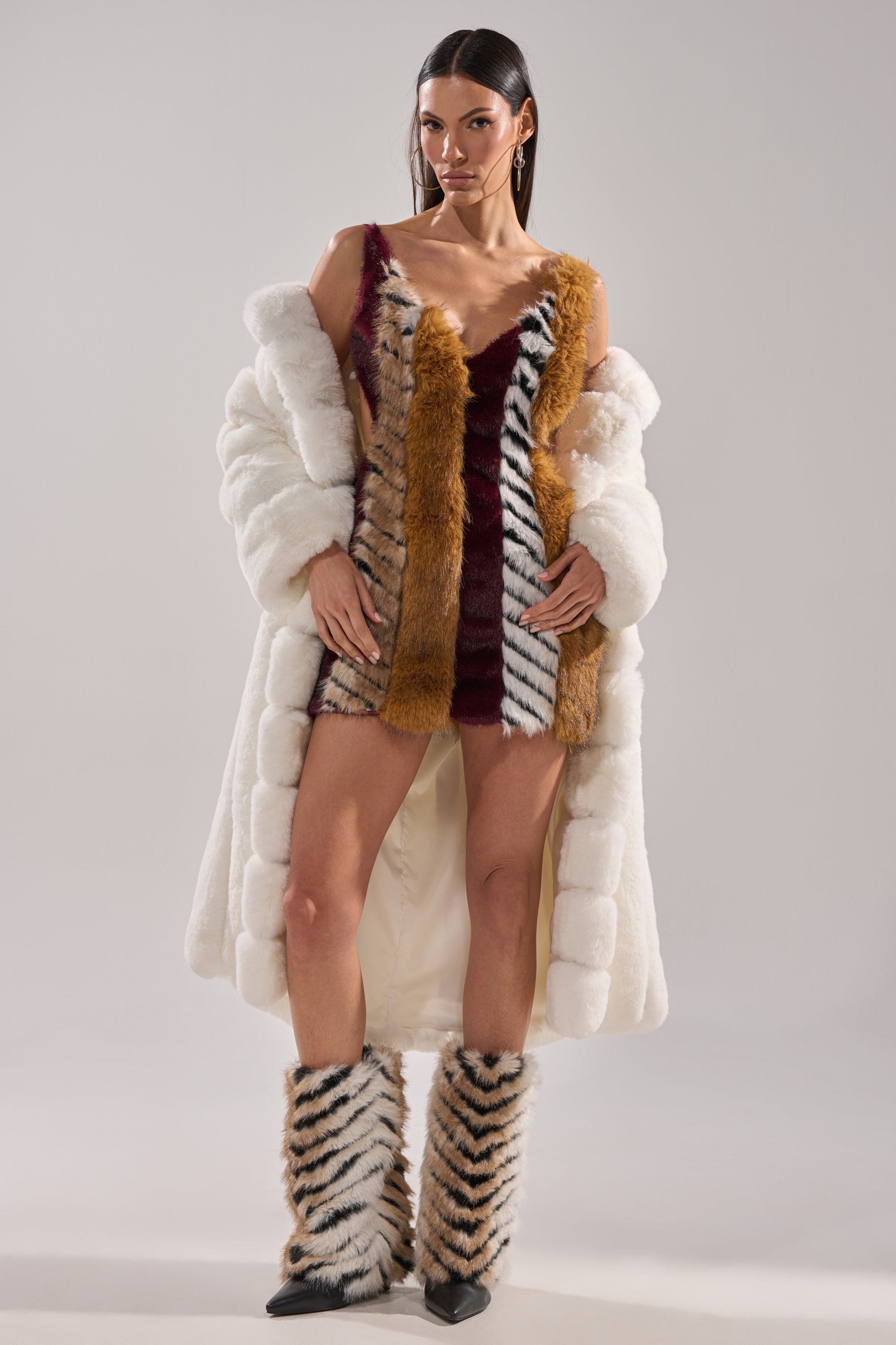 A woman confidently wears the ANIMALISTIC MINI DRESS with a white faux fur coat and matching tiger-stripe boots featuring satin lining, posing against a plain background.