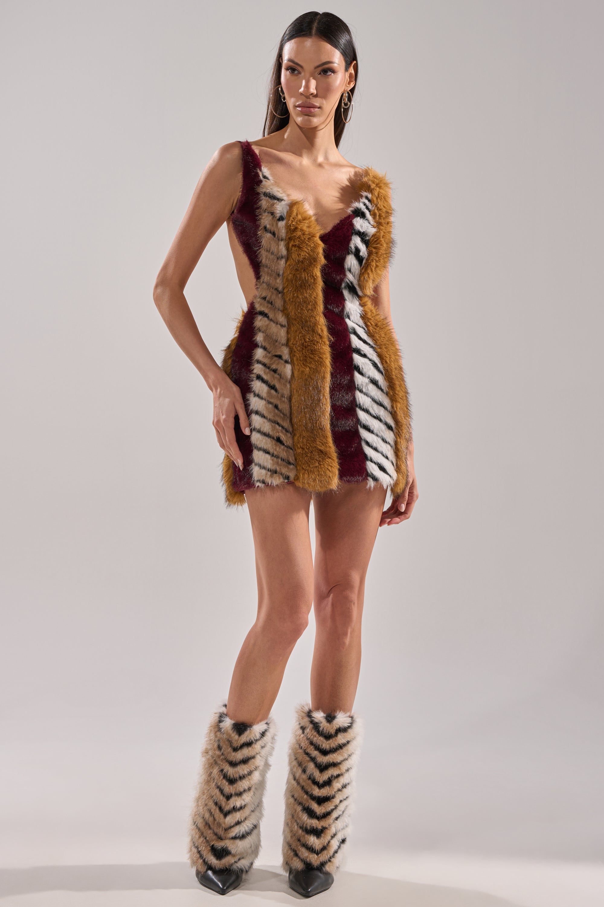A woman stands wearing the ANIMALISTIC MINI DRESS, featuring a deep V-neck and colorblock faux fur panels in brown, burgundy, and white with black stripes. She completes the look with matching faux fur boots and a satin lining.