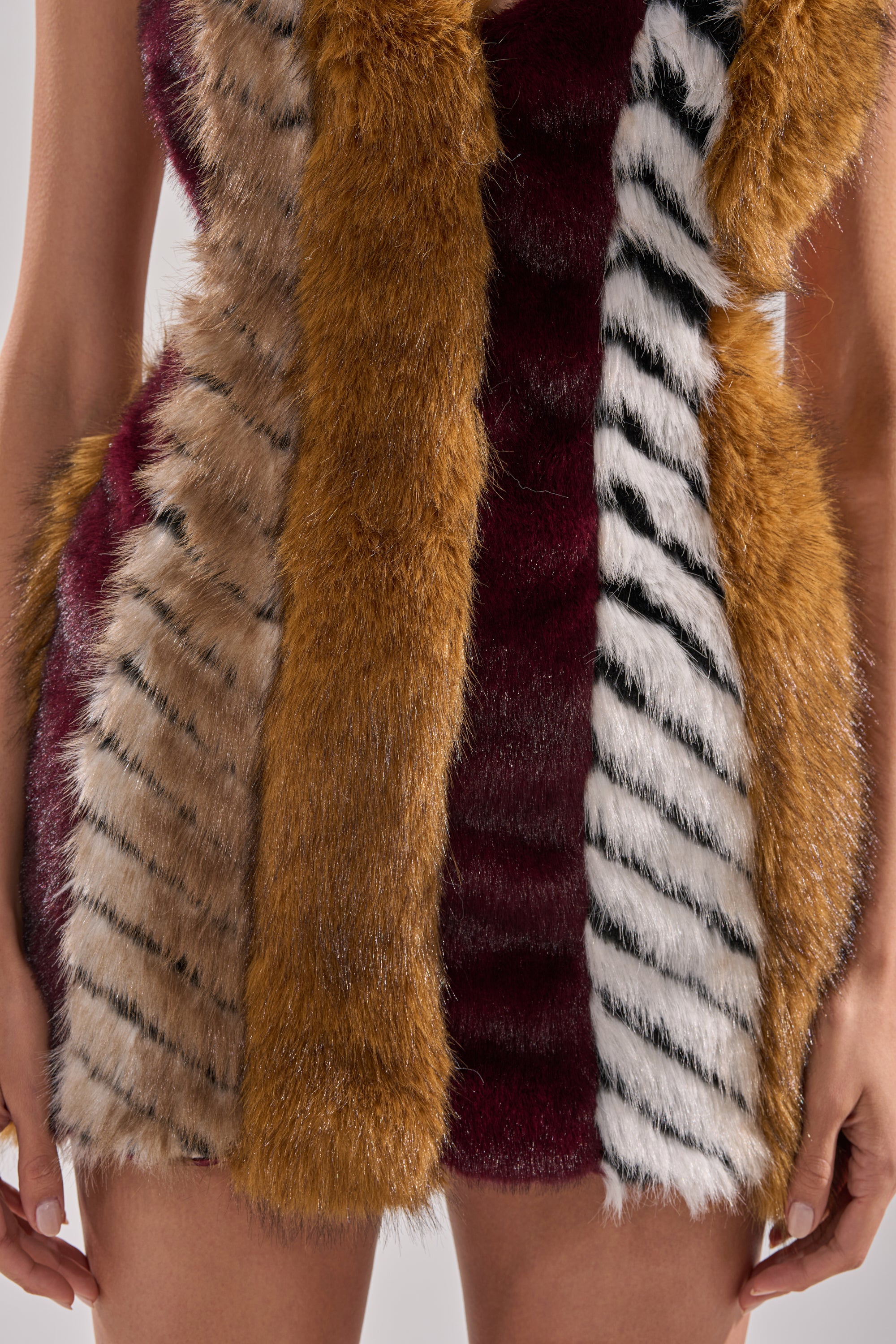 A close-up of someone wearing the ANIMALISTIC MINI DRESS, made of striped faux fur in brown, black, white, and burgundy with satin lining, their arms visible at their sides.