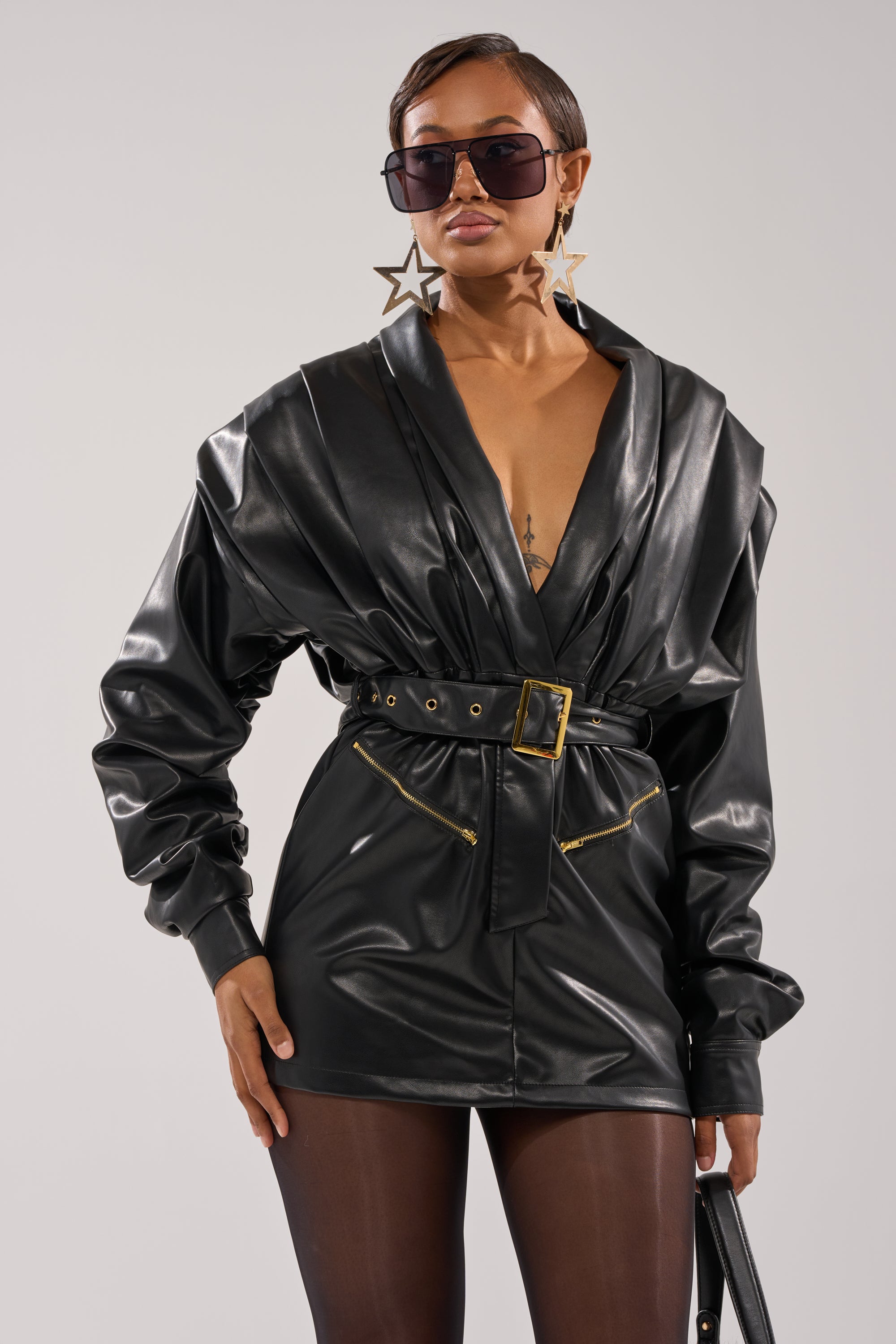 Wearing the RIDE OR DIE FAUX LEATHER MINI DRESS, sheer black tights, star-shaped earrings, and oversized sunglasses, a confident individual stands against a plain background.