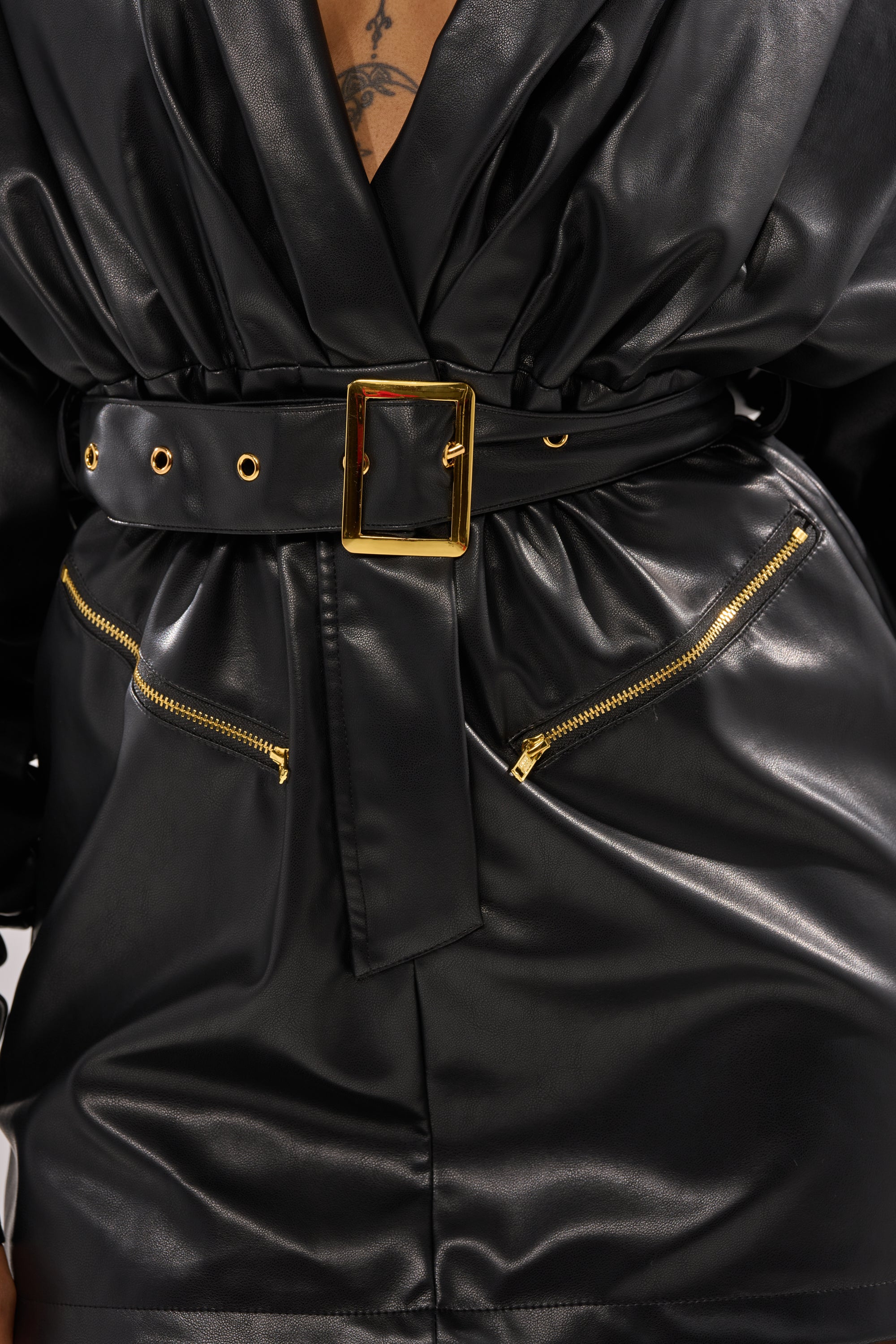 A close-up of a person wearing the RIDE OR DIE FAUX LEATHER MINI DRESS in black, featuring a plunging neckline, wide gold-buckled belt, and two gold-zippered pockets. A portion of a chest tattoo is visible above the neckline.