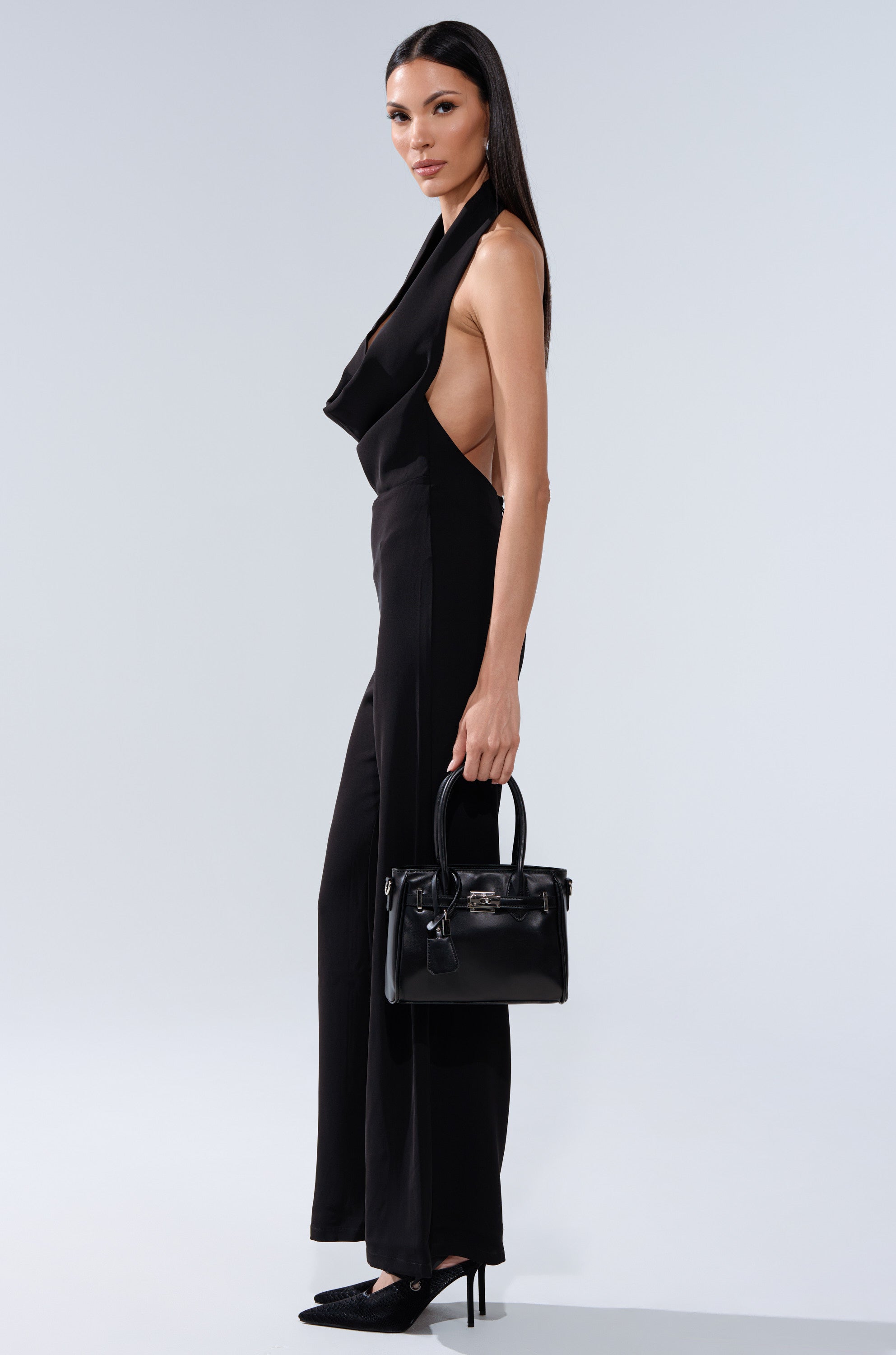 A woman stands against a light gray background wearing the SLEEK AND CHIC JUMPSUIT—a sleeveless black jumpsuit with a cowl neckline. She holds a black handbag, wears black high heels, and has long straight hair.