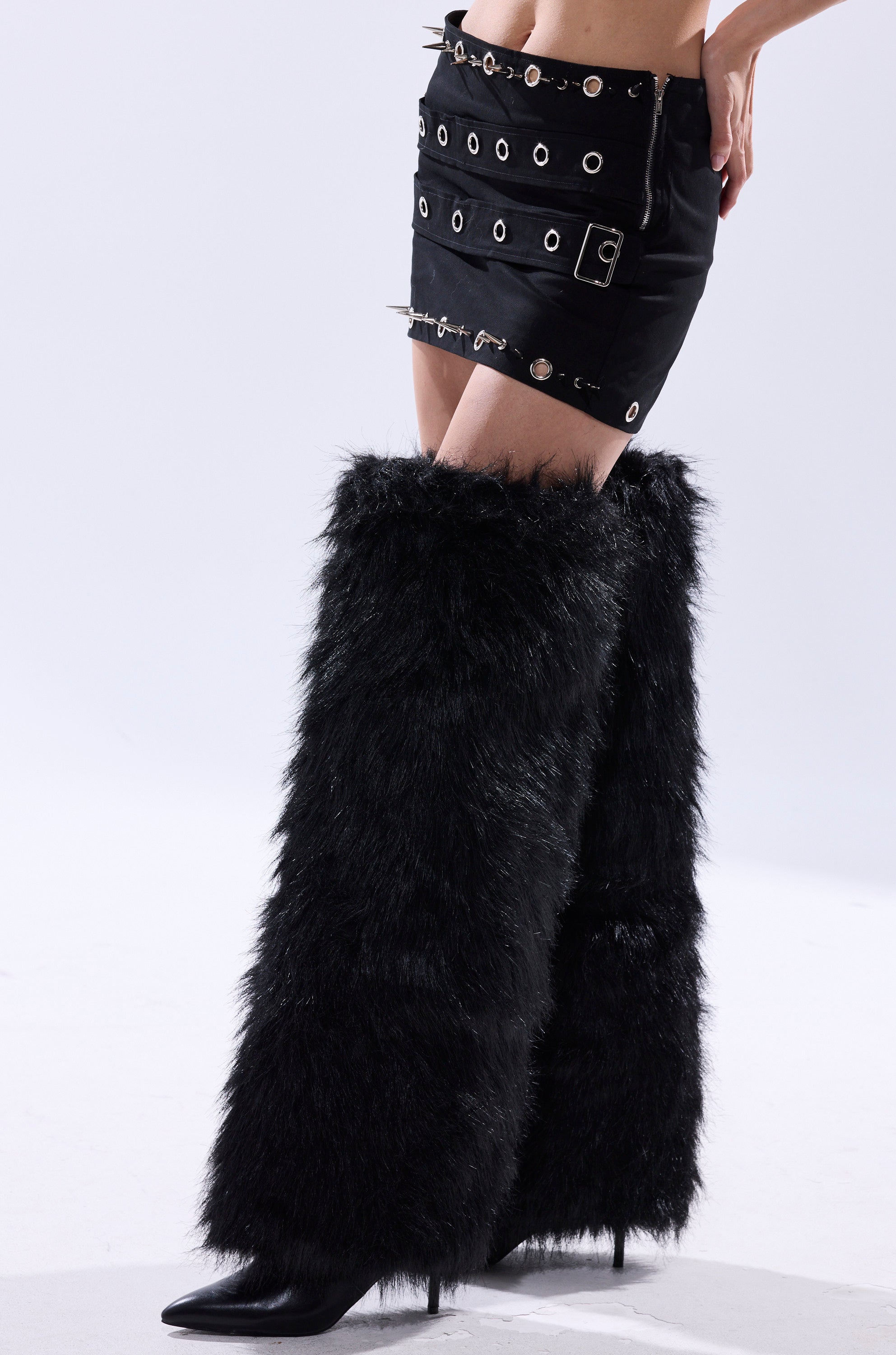 A person wearing the CRAZY FOR YOU MINI SKIRT with metal eyelet details and black furry leg warmers over high-heeled boots, posing against a plain light background.