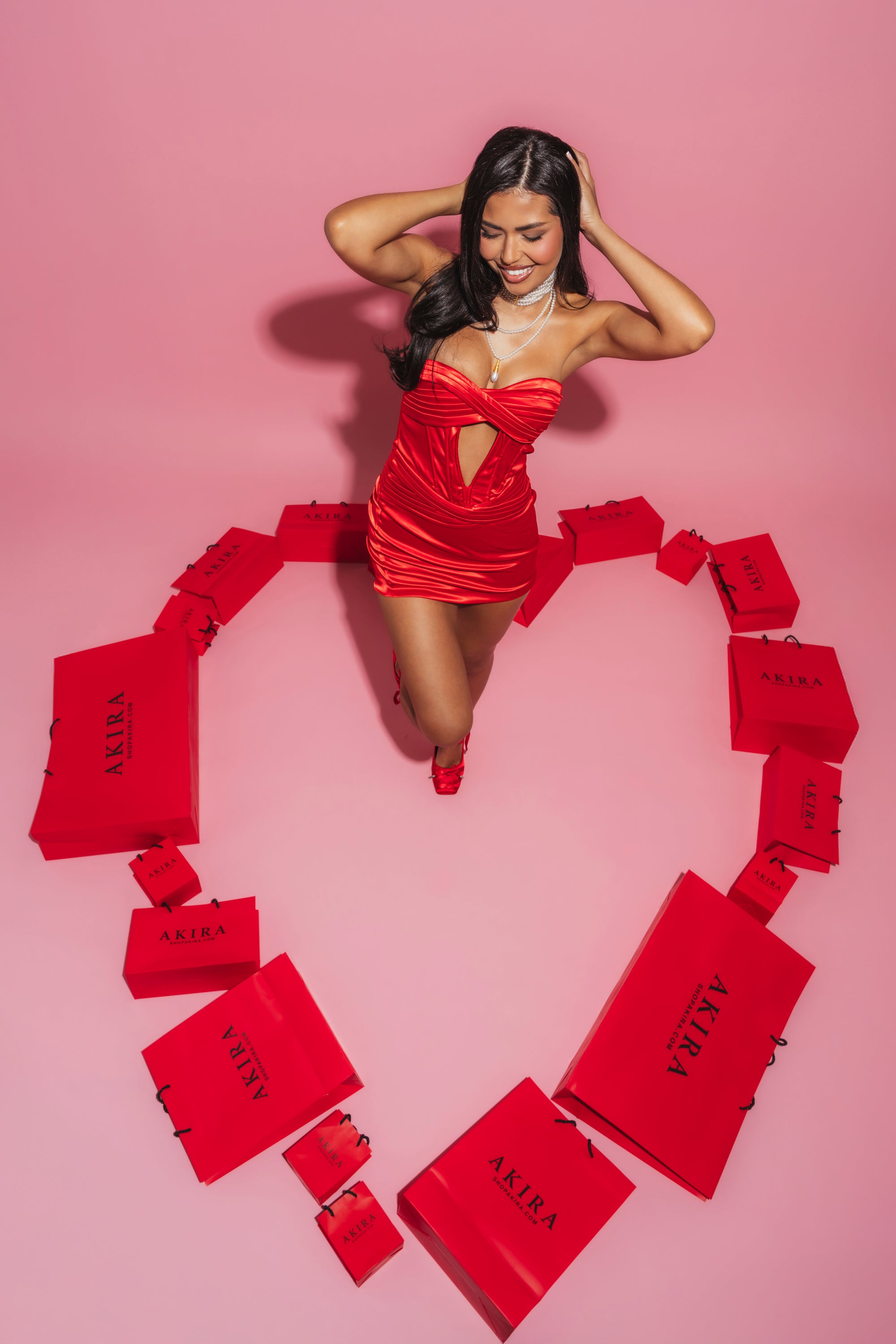 A woman in the V DAY CUPIDS CRUSH MINI DRESS, a red satin mini with a corset-inspired bodice, stands beside a heart made of shopping bags. 5