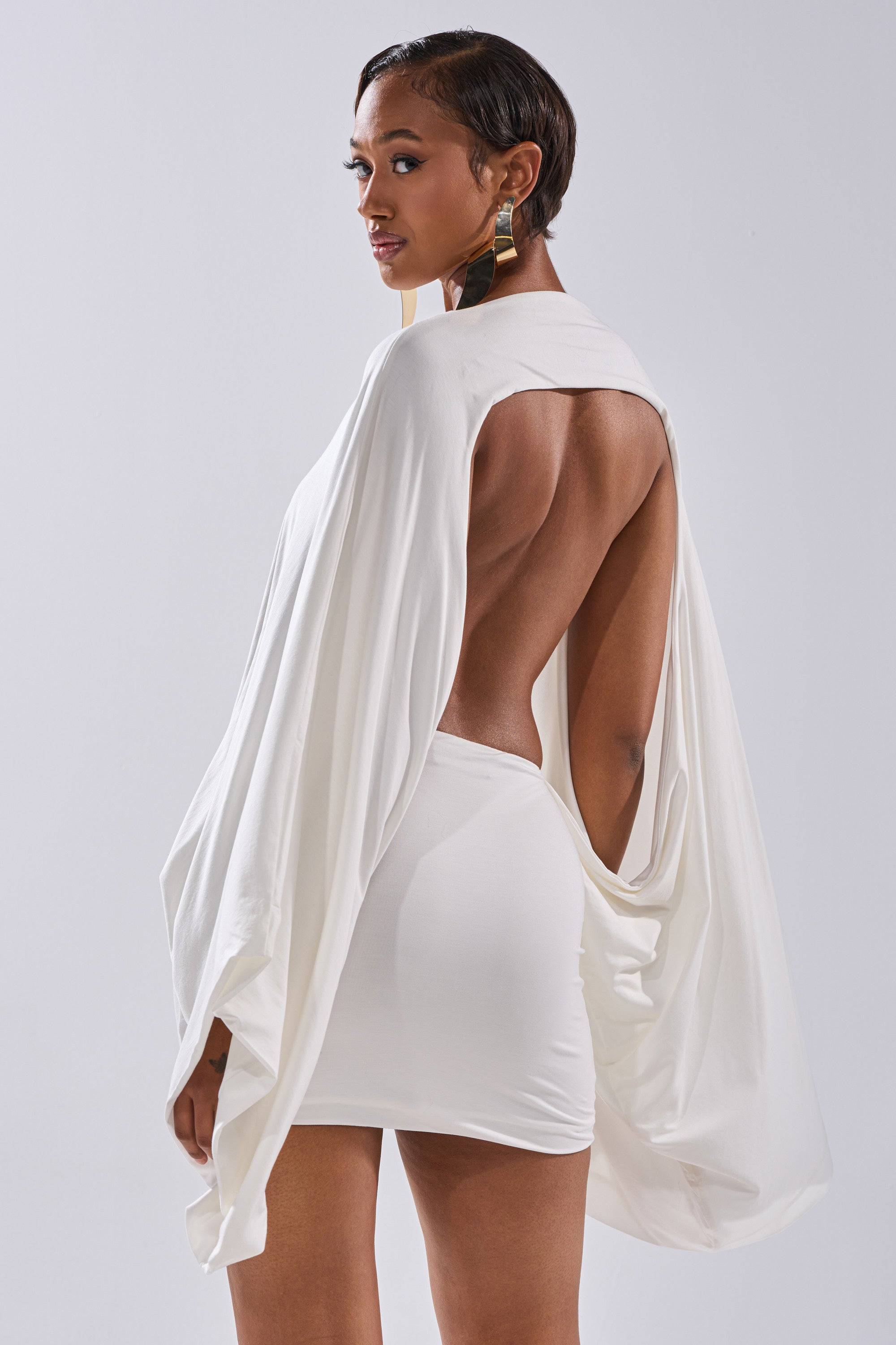 A woman with short hair and statement earrings poses in the DIANA DRAPED MINI DRESS IN WHITE, featuring an open back and flowing draped sleeves, against a plain light background.