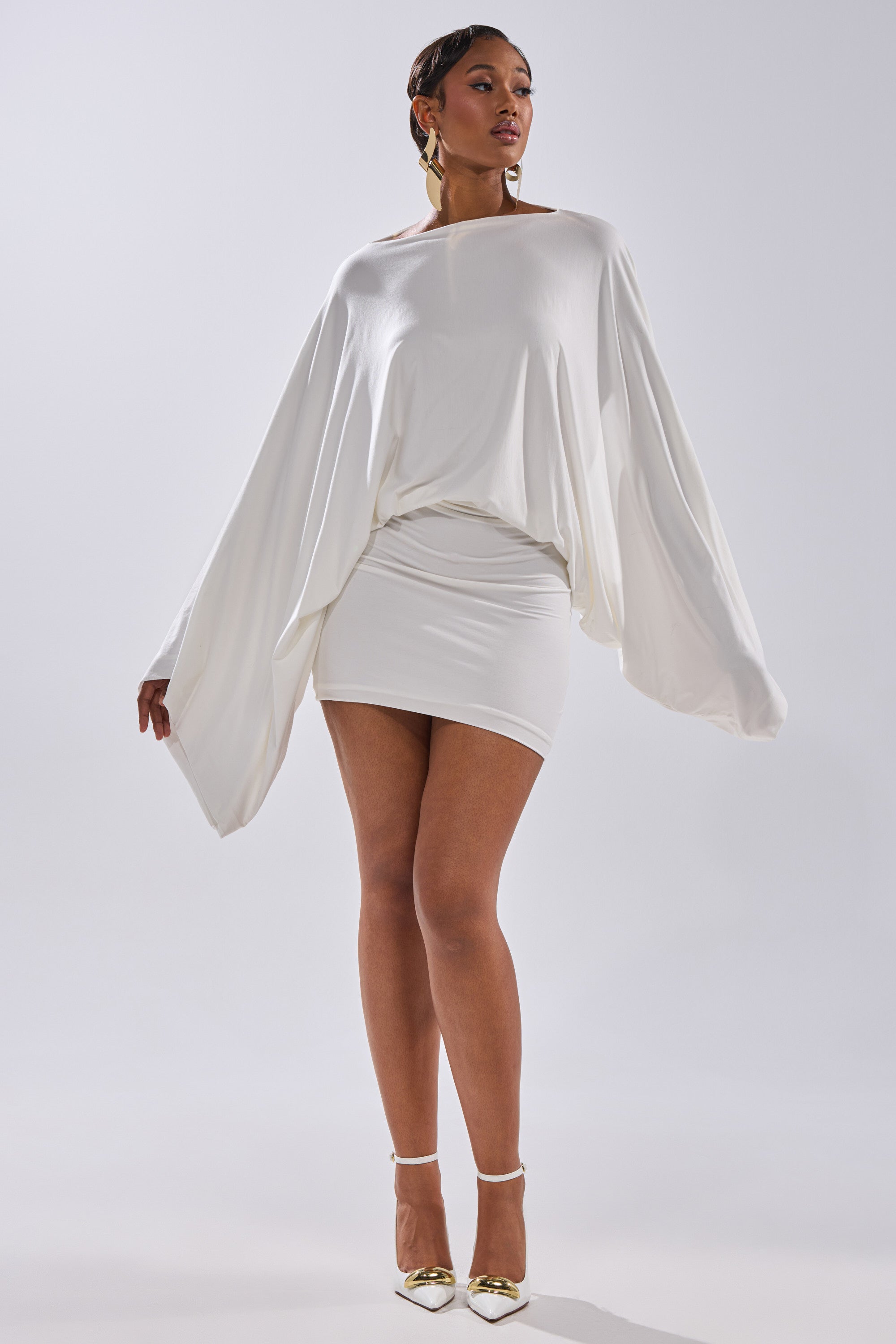 A woman exudes confidence in the DIANA DRAPED MINI DRESS IN WHITE, featuring wide batwing sleeves and a fitted mini skirt. She styles it with short hair, gold earrings, and gold heels against a plain light background.