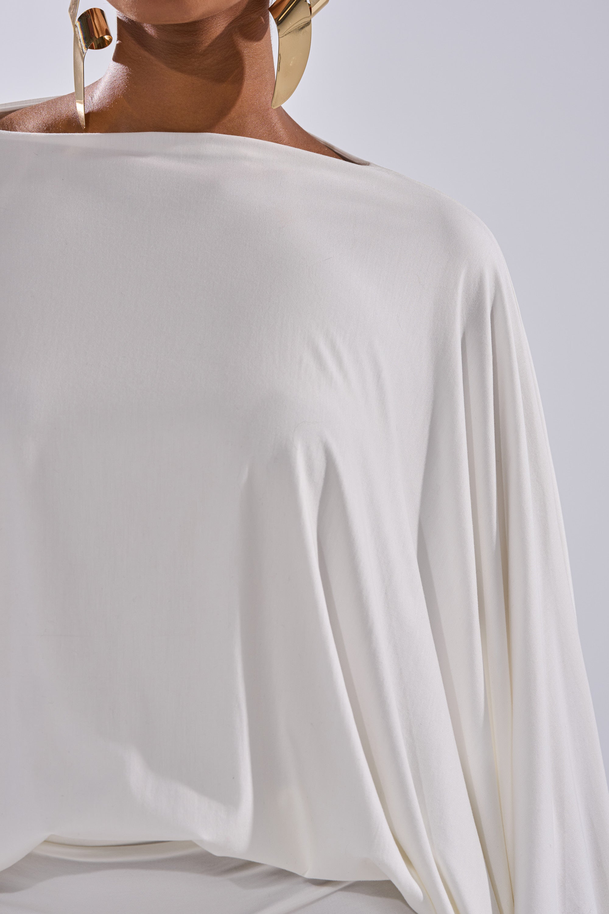 A person wears the DIANA DRAPED MINI DRESS IN WHITE with large gold statement earrings, photographed from shoulders to waist against a light background.