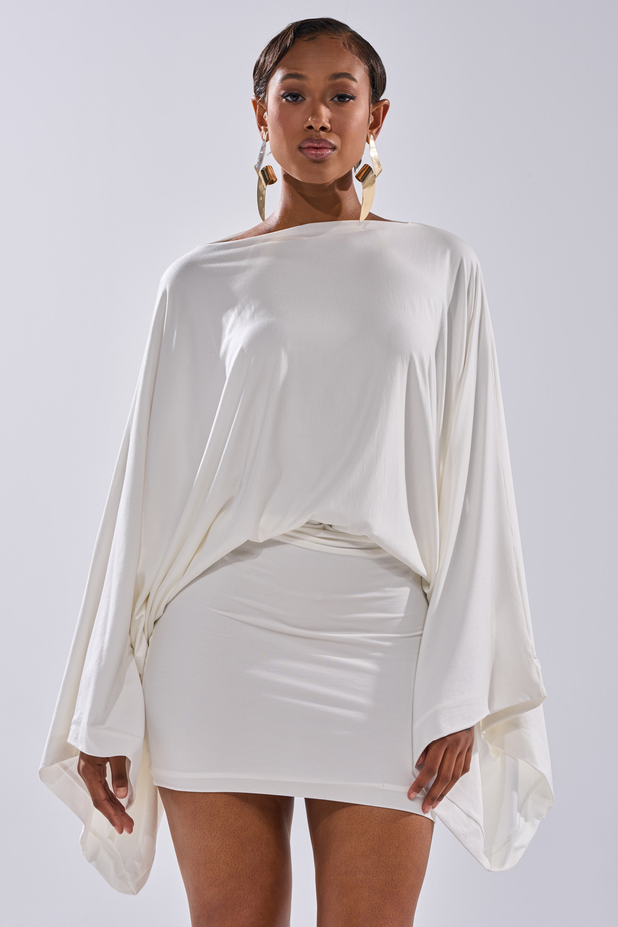 A woman stands facing the camera against a plain light background, wearing the DIANA DRAPED MINI DRESS IN WHITE with dramatic batwing sleeves and a fitted skirt, styled with large geometric gold earrings.