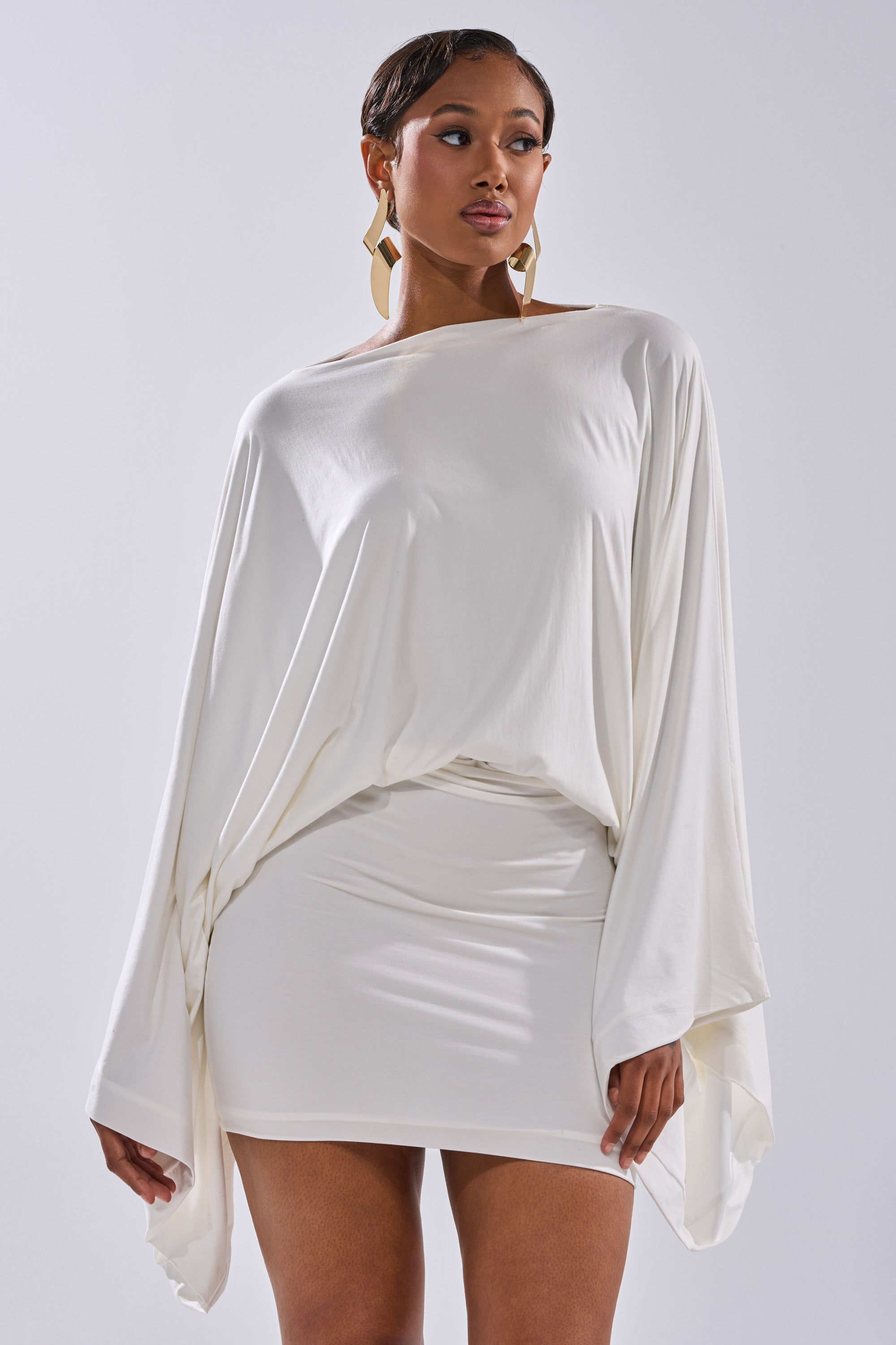 A woman wears the DIANA DRAPED MINI DRESS IN WHITE featuring a loose, long-sleeved top and fitted skirt. She has short hair, bold geometric earrings, and stands against a plain light background.