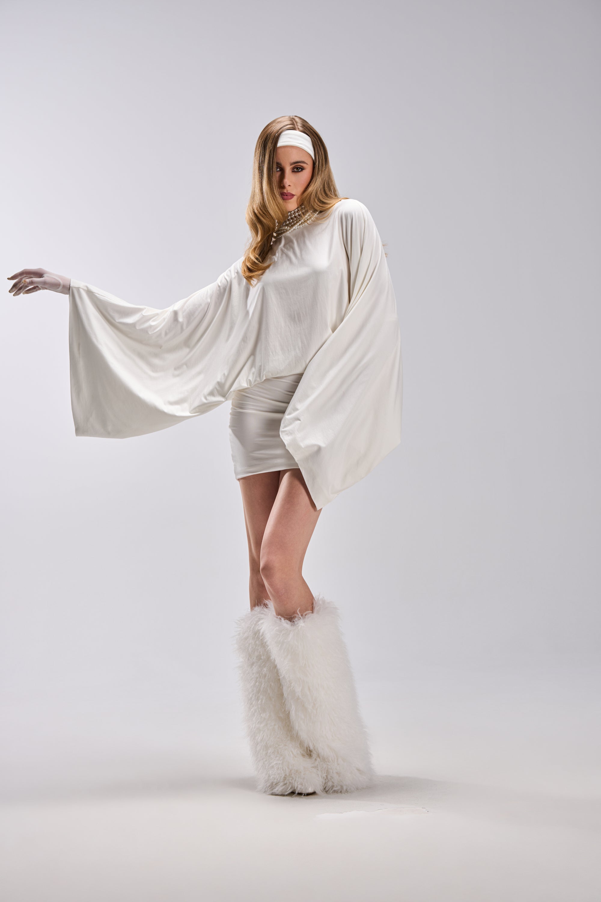 A woman poses against a plain light gray background wearing the DIANA DRAPED MINI DRESS IN WHITE, featuring wide, flowing sleeves and a short skirt, styled with a headband and tall, fluffy white boots.