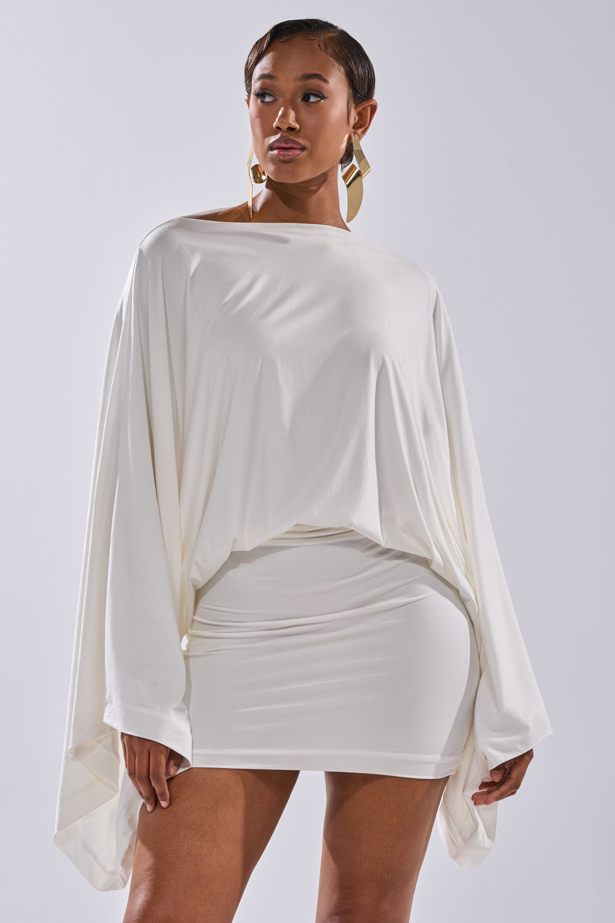 A woman with short hair and gold geometric earrings stands confidently in the DIANA DRAPED MINI DRESS IN WHITE, featuring long sleeves and a fitted mini skirt, looking to the side against a plain background.