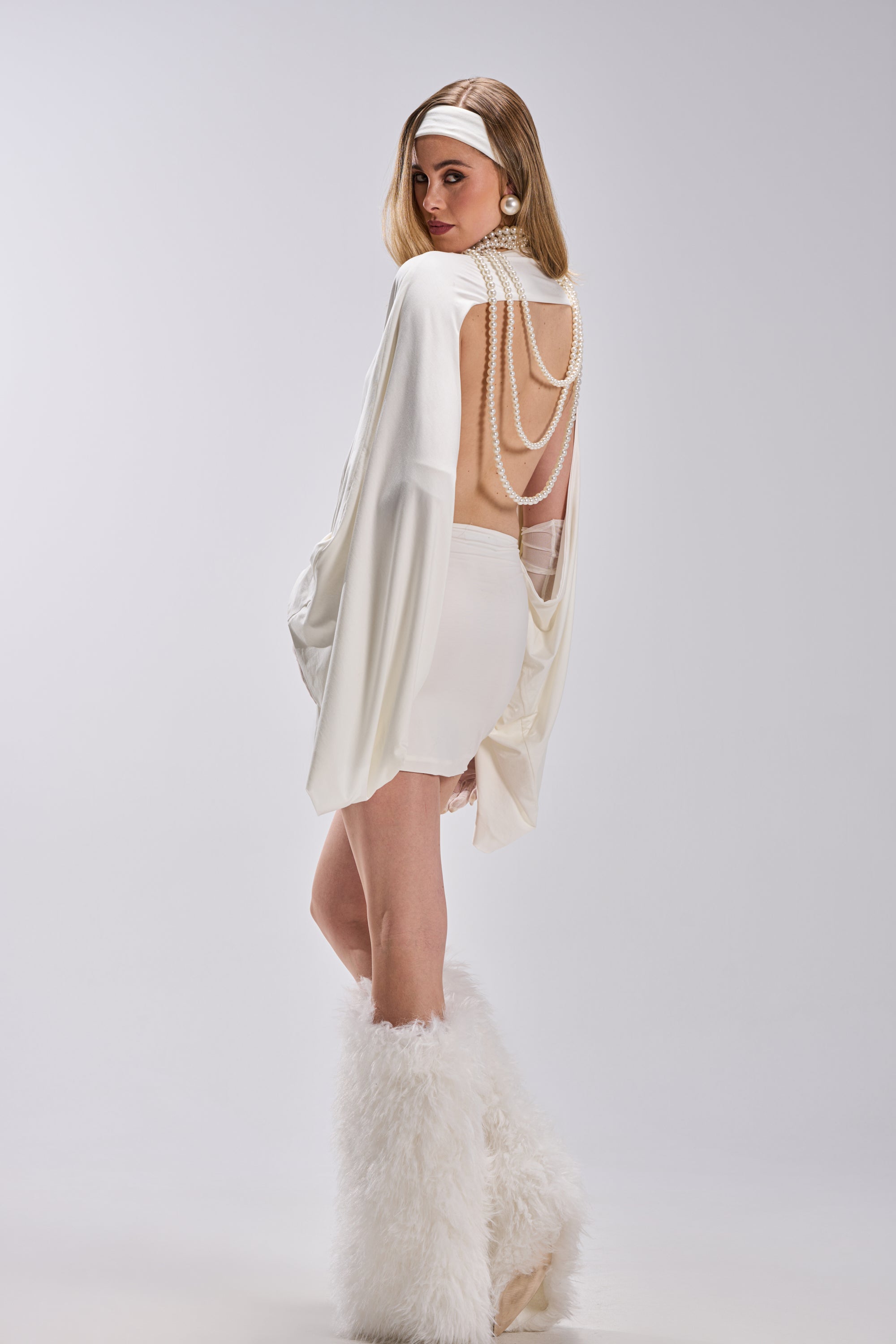 A woman models the DIANA DRAPED MINI DRESS IN WHITE, styled with pearl chains, a white headband, statement earrings, and knee-high furry boots—creating a stunning outfit ideal for special occasions.