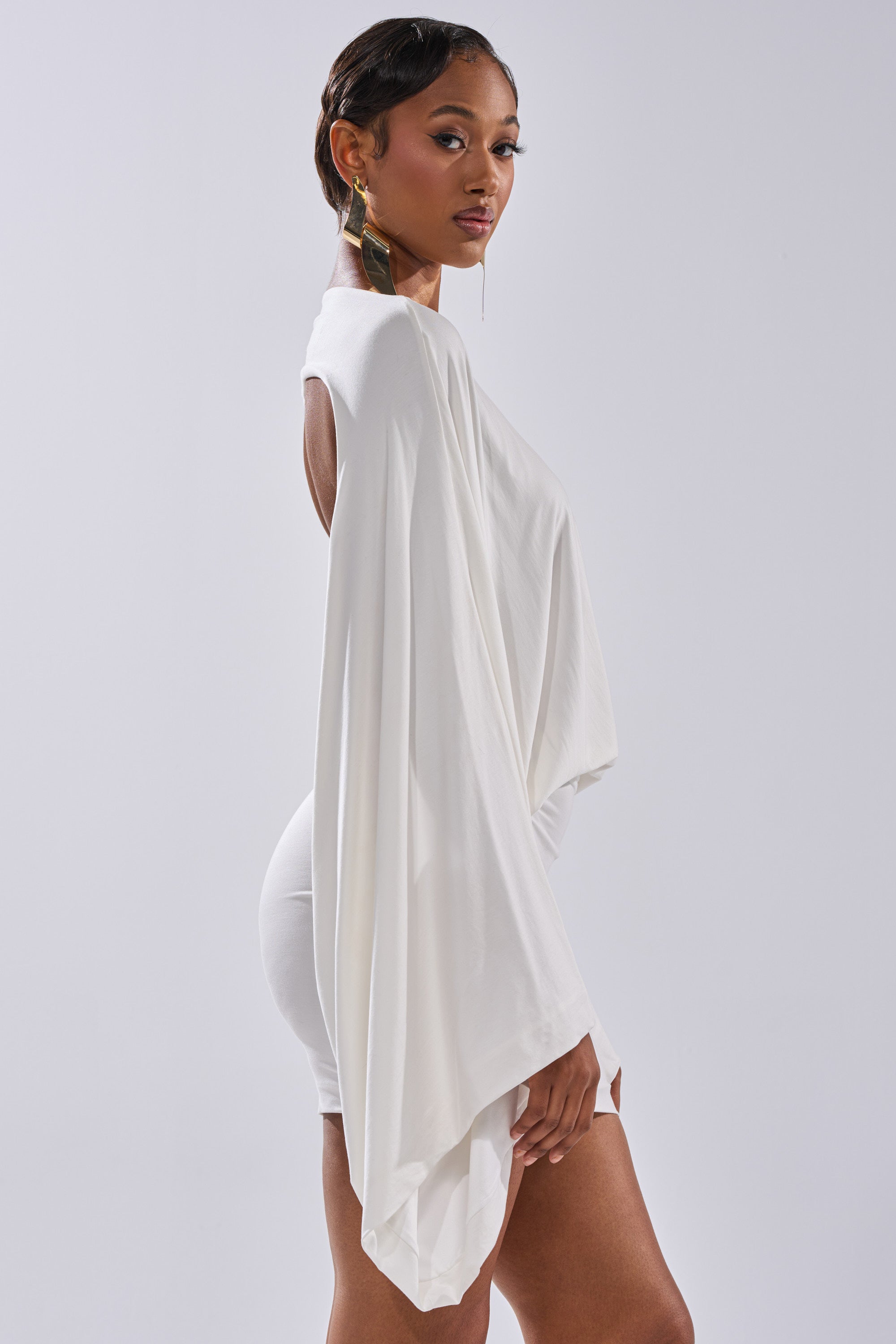 A woman wearing the DIANA DRAPED MINI DRESS IN WHITE stands sideways, gazing confidently at the camera. She has short, styled hair and wears long geometric gold earrings against a plain light background.