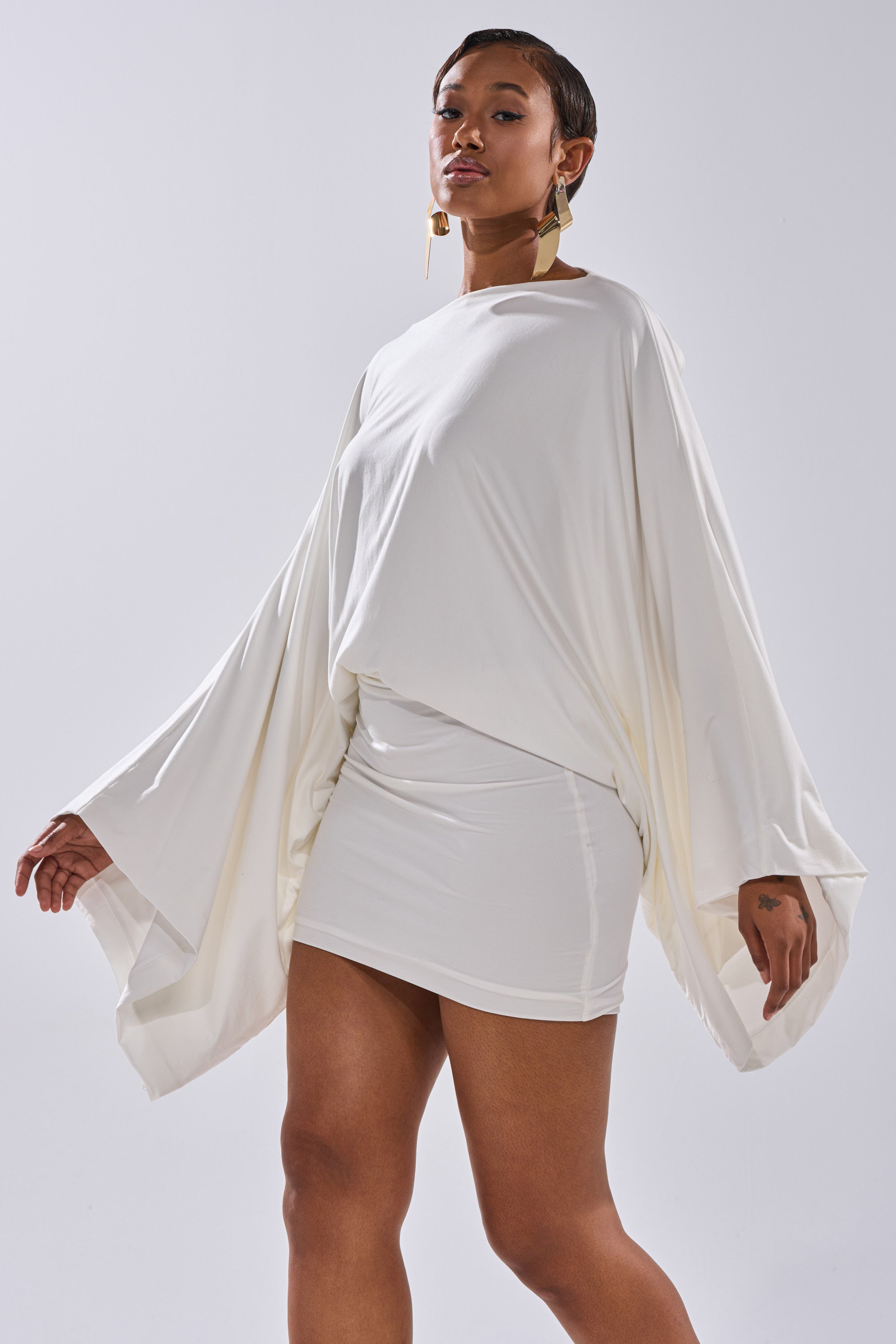 A woman poses confidently in the DIANA DRAPED MINI DRESS IN WHITE, a loose mini dress with wide, flowing sleeves and a fitted skirt, accessorized with large gold earrings and a choker against a plain, light background.