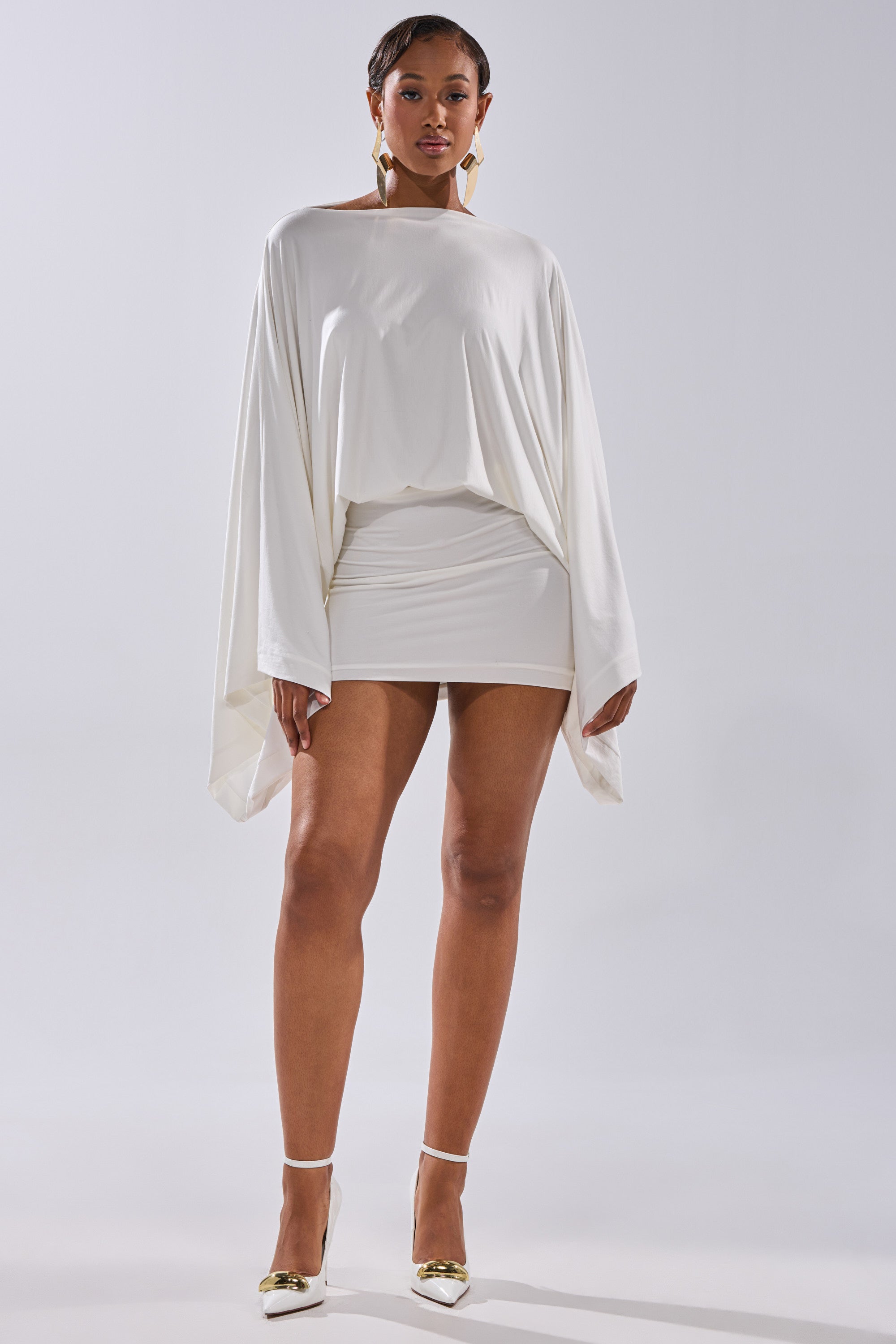A woman stands confidently in the DIANA DRAPED MINI DRESS IN WHITE, featuring batwing sleeves, paired with gold and white heels and statement earrings, set against a plain light background.